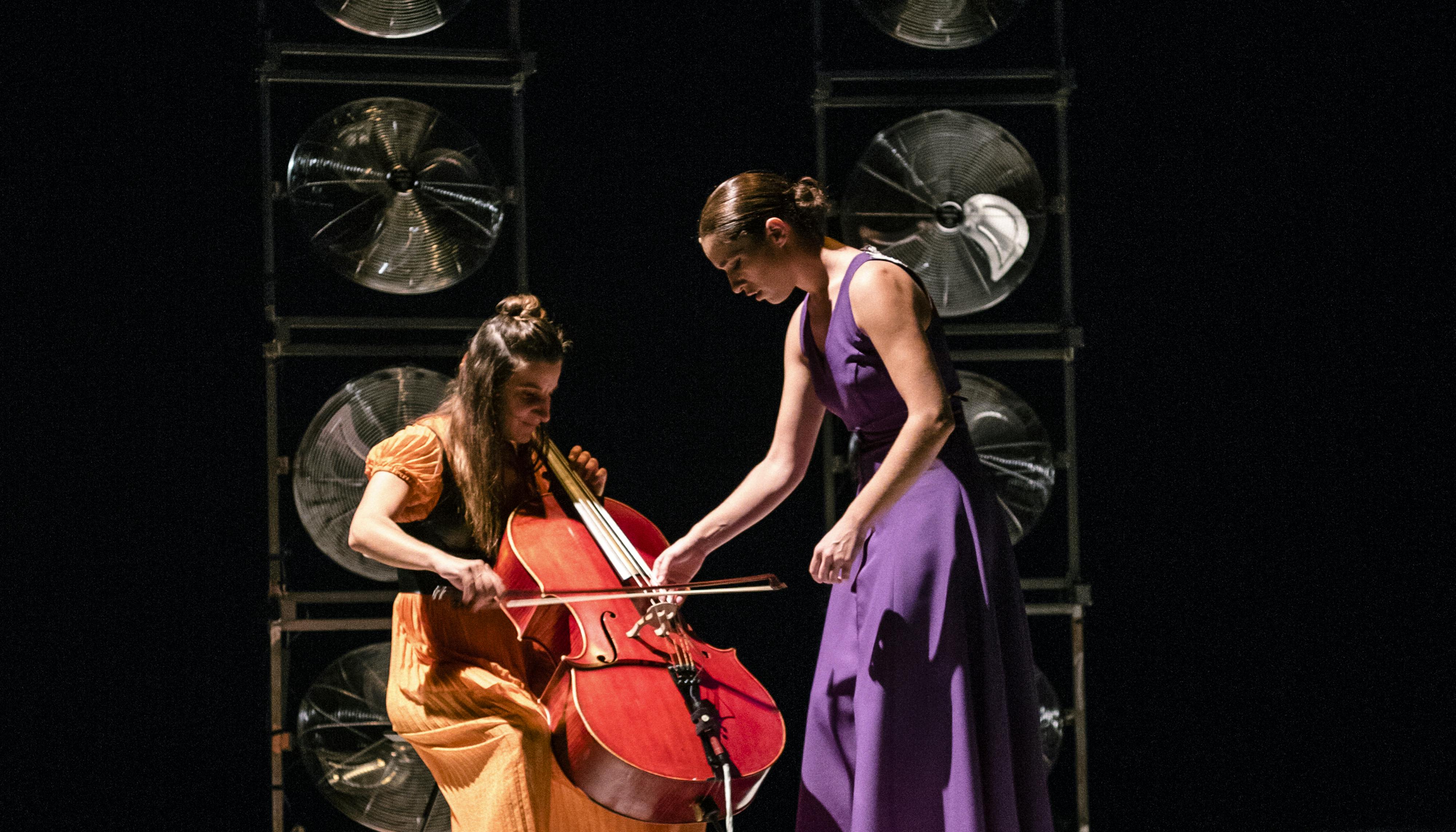 Two performers on stage. One of them, standing, wearing a long purple dress, bends over the cello of the second one, in a long orange dress, who is playing it.