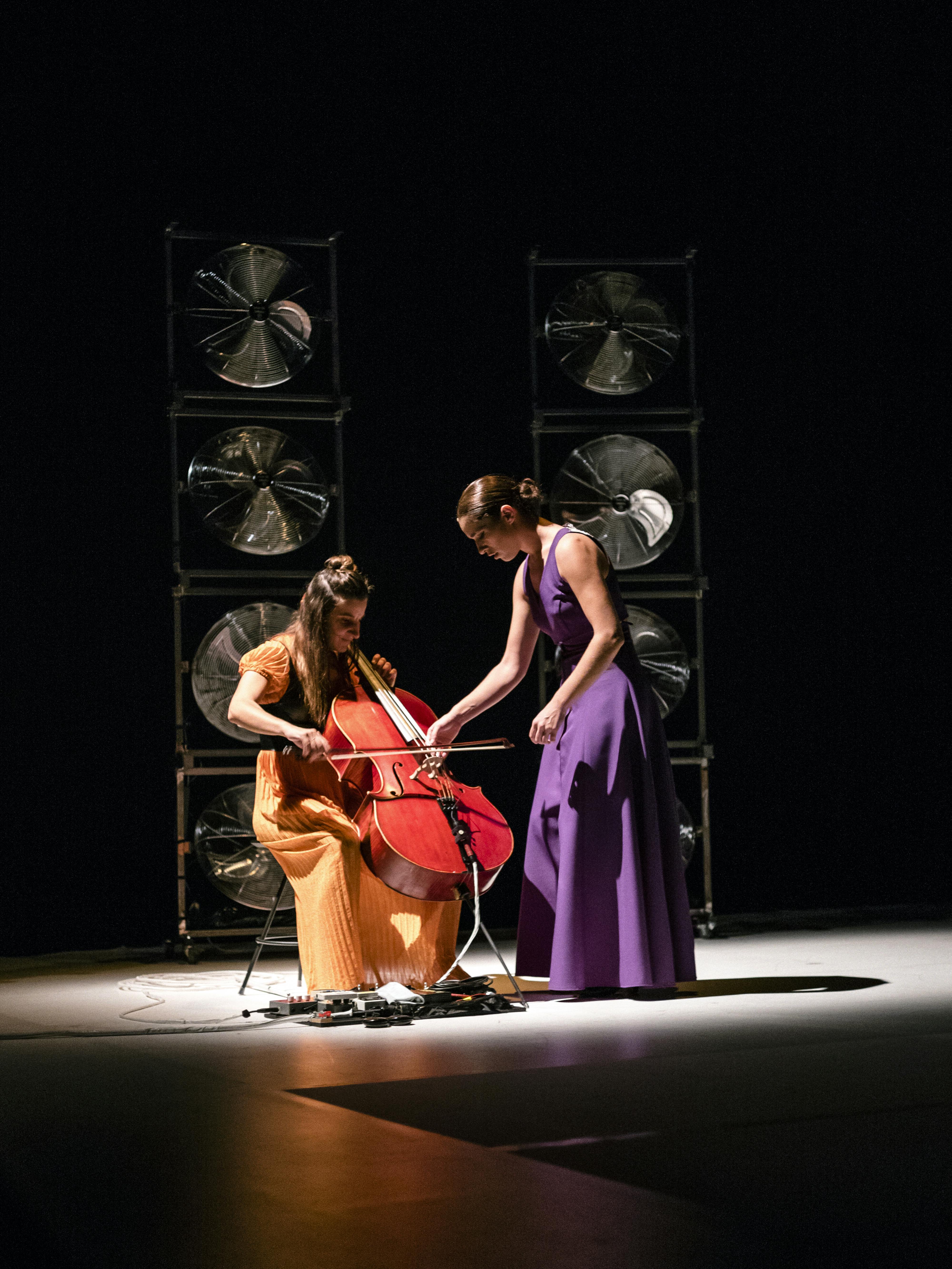 Due performer sulla scena. Una delle due, in piedi, con indosso un lungo abito viola, si china sul violoncello della seconda, in abito arancione, intenta a suonarlo.