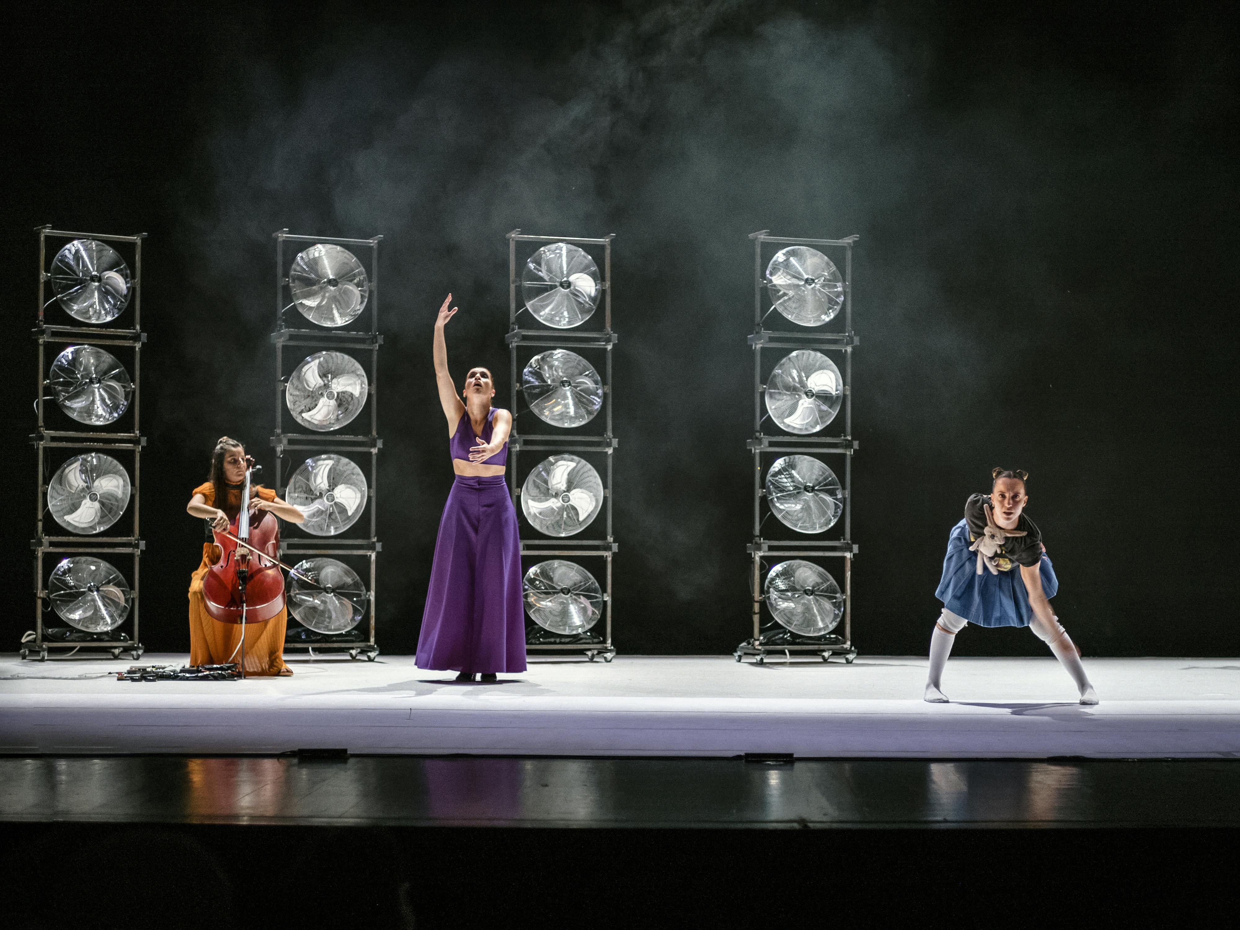Le tre performer di YouMe, una violoncellista, una danzatrice di flamenco e una proveniente dall'hip hop, durante la performance. Dietro di loro quattro colonne di ventilatori impilati tra loro e del fumo scenico sullo sfondo buio.