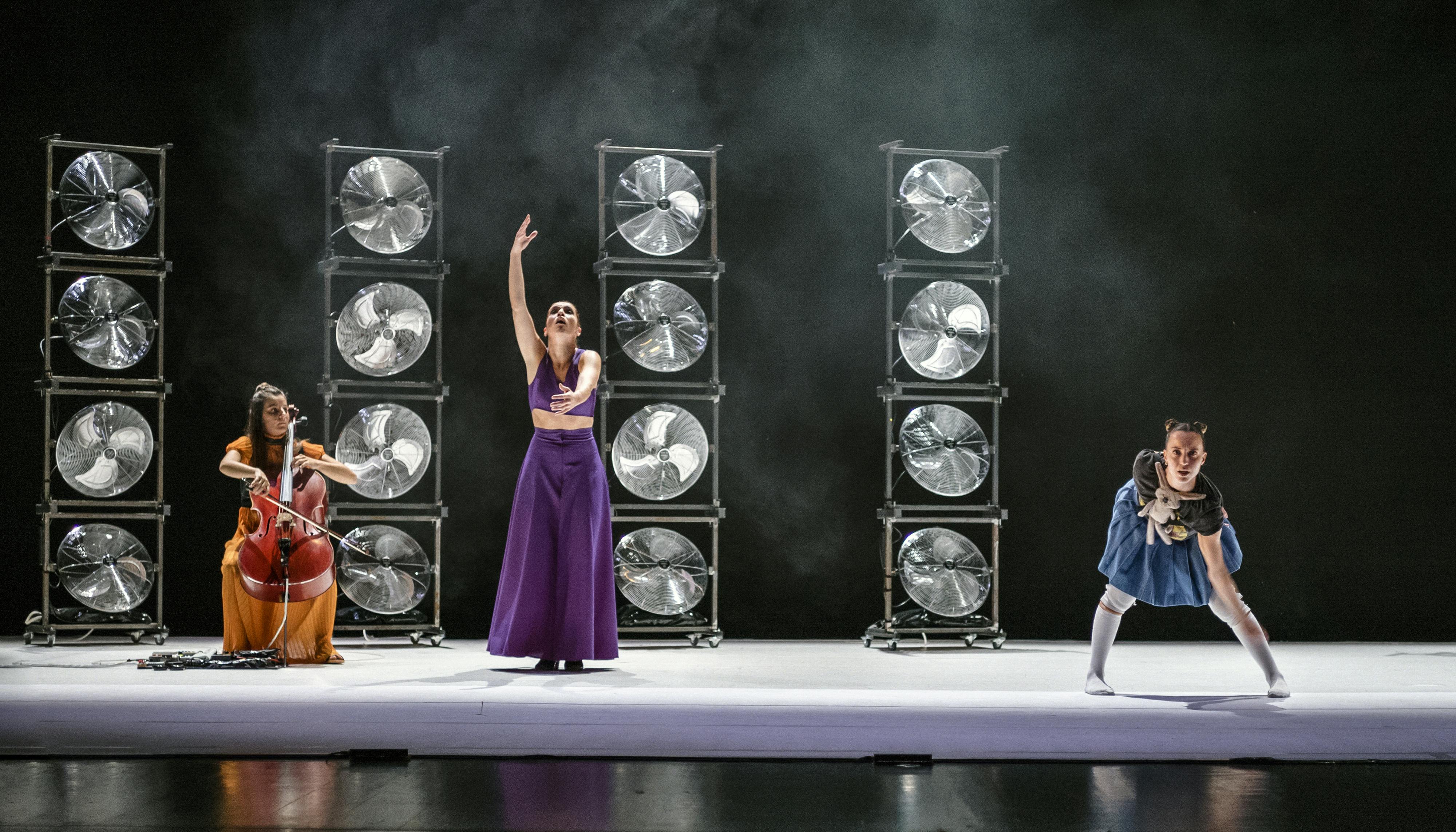 The three YouMe performers (a cellist, a flamenco dancer and a hip hop dancer) during the performance. Behind them four pillars made of fans stacked together and some stage smoke in the dark background.