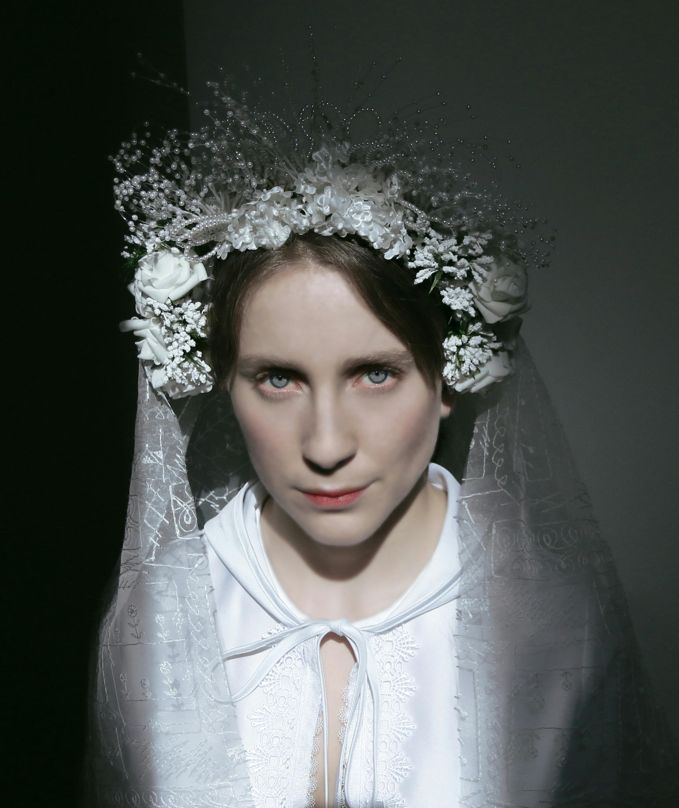 Half-length portrait of a woman in a soft white dress. On her head she wears a wreath of flowers, also white, from which descends a long elaborate veil.