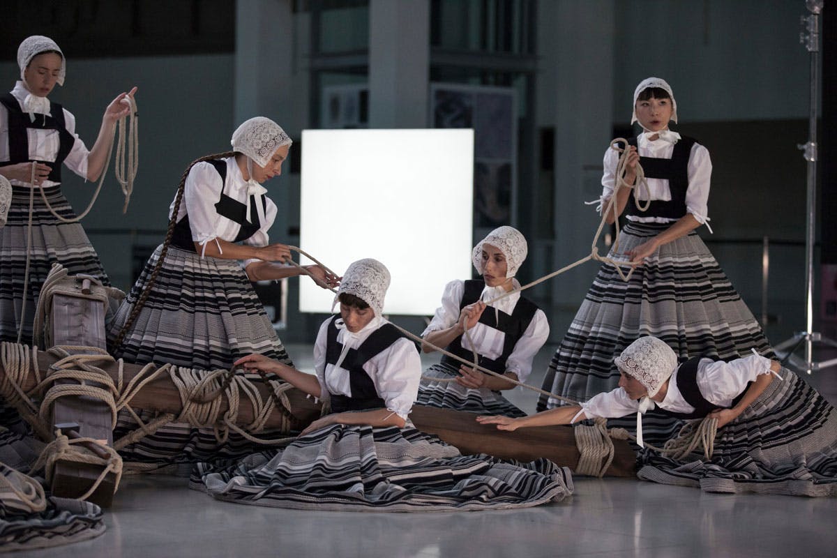 Photograph of the show "Sonoma": some women, in traditional clothes with wide skirts, hold and pull ropes tied around a large wooden crucifix lying on the stage.