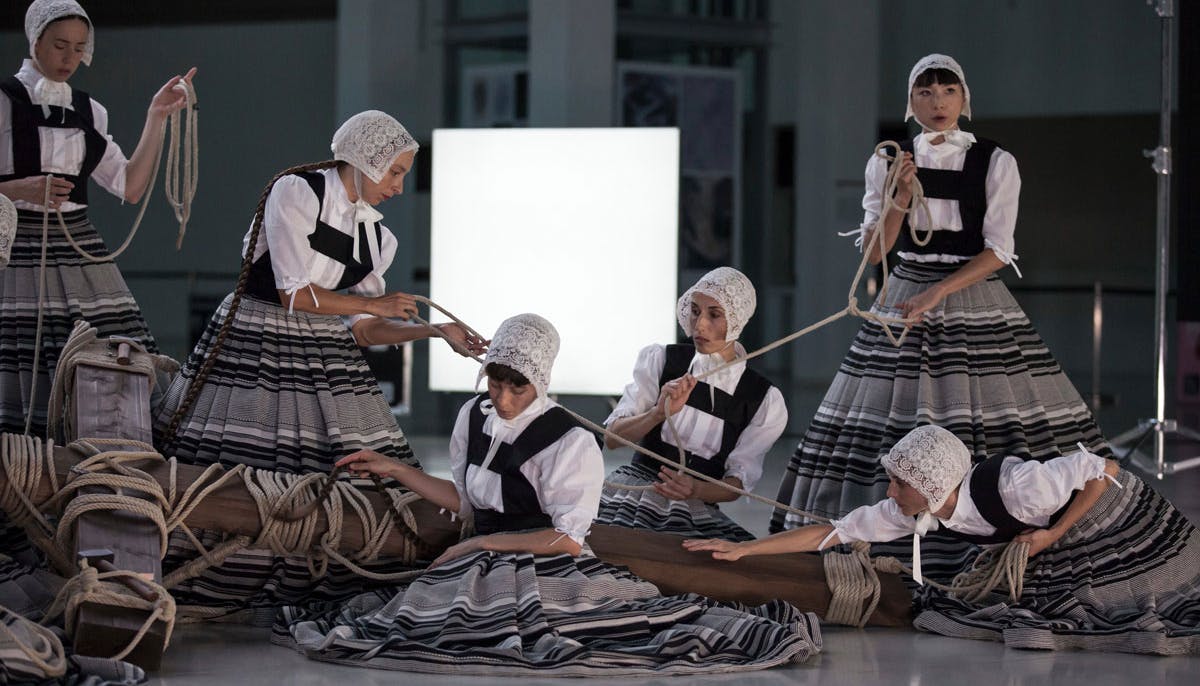Photograph of the show "Sonoma": some women, in traditional clothes with wide skirts, hold and pull ropes tied around a large wooden crucifix lying on the stage.