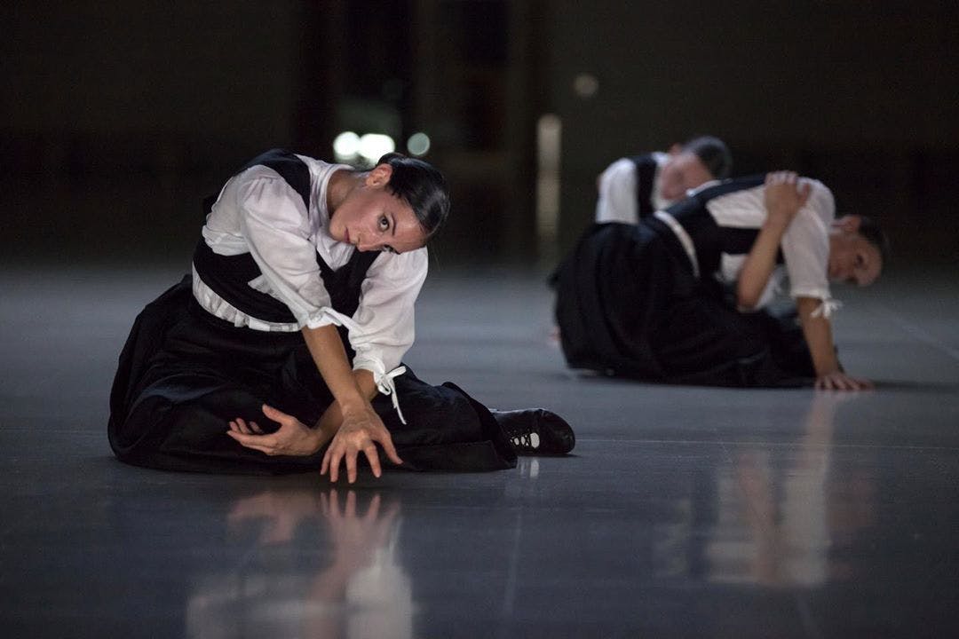 Three dancers on the stage during the performance Sonoma. They are sitting on the floor with their legs bent on one side of their bodies and their arms crossed in front of them.