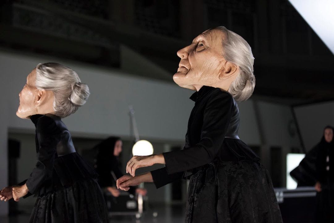 Photograph of the show Sonoma: in the foreground we see two bodies wearing large masks representing elderly women with long white hair pulled back.