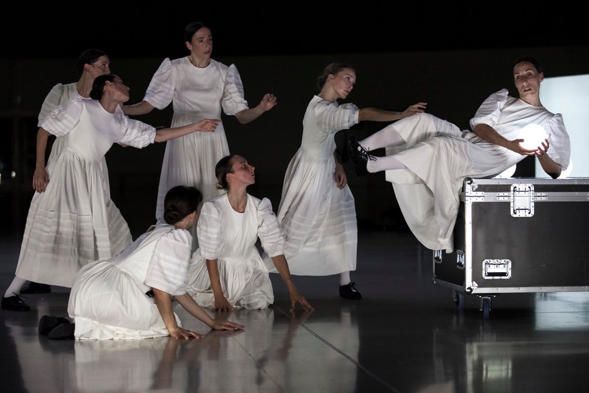 Photograph of the Sonoma show: some dancers, in white traditional short-sleeved dresses, reach out to a companion. She is suspended above a cage and is holding a glowing sphere.