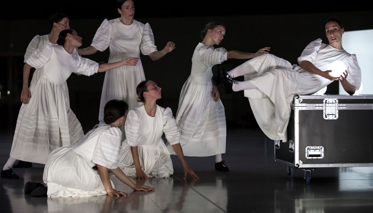 Fotografia dello spettacolo Sonoma: alcune danzatrici, in bianchi abiti tradizionali a maniche corte, si protraggono verso una compagna sospesa sopra un baule su ruote che tiene in mano una sfera luminosa.