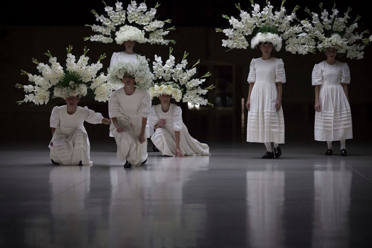 Fotografia dello spettacolo Sonoma: alcune donne, in bianchi abiti tradizionali senza maniche, guardano verso la camera. Sul capo indossano grandi corone di fiori che incorniciano i loro volti come aureole.