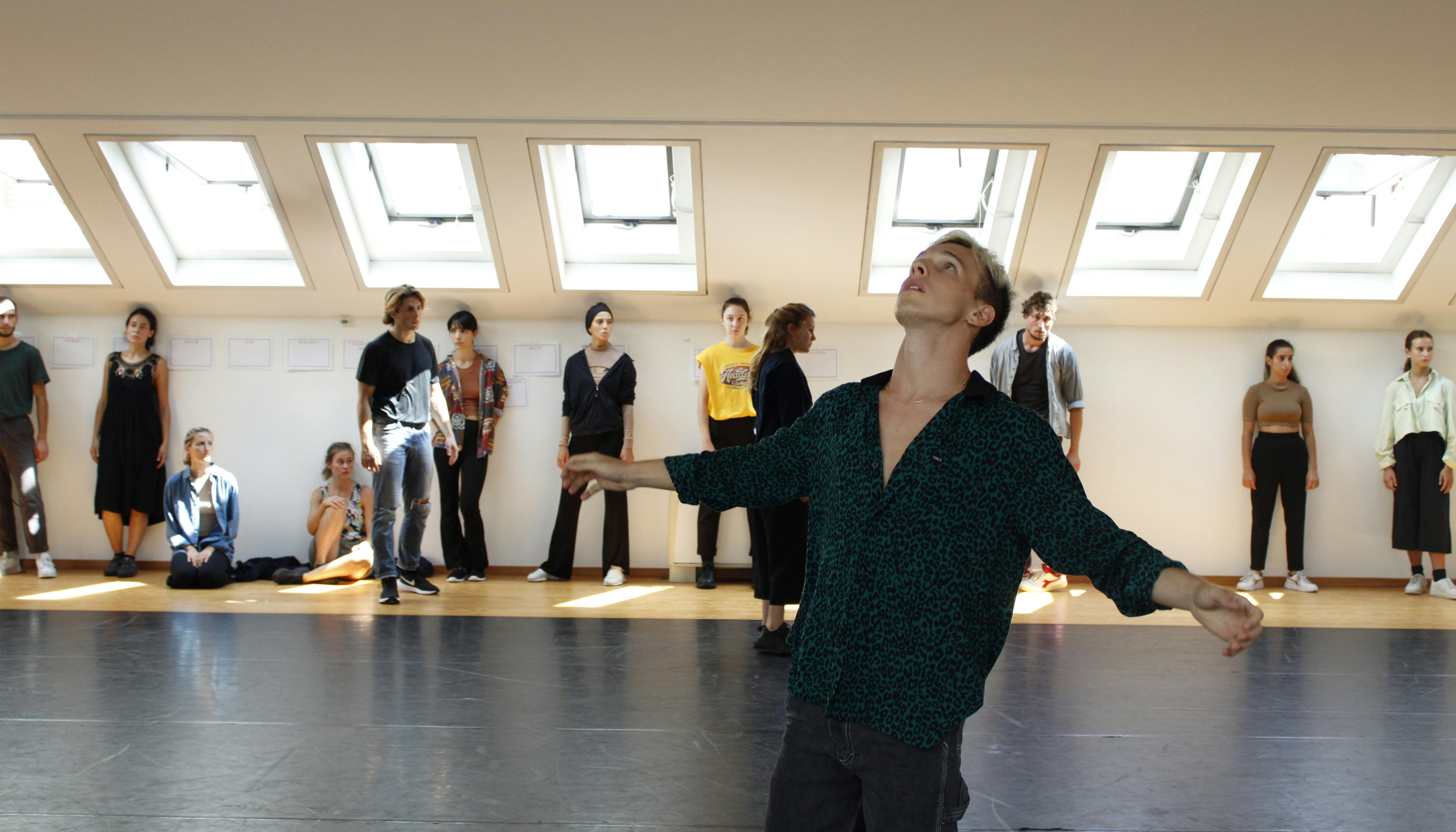 Un danzatore, in piedi al centro dello Studio, apre le braccia guardando verso l'alto. A fondo sala un gruppo di performer, appoggiati alla parete, osservano.