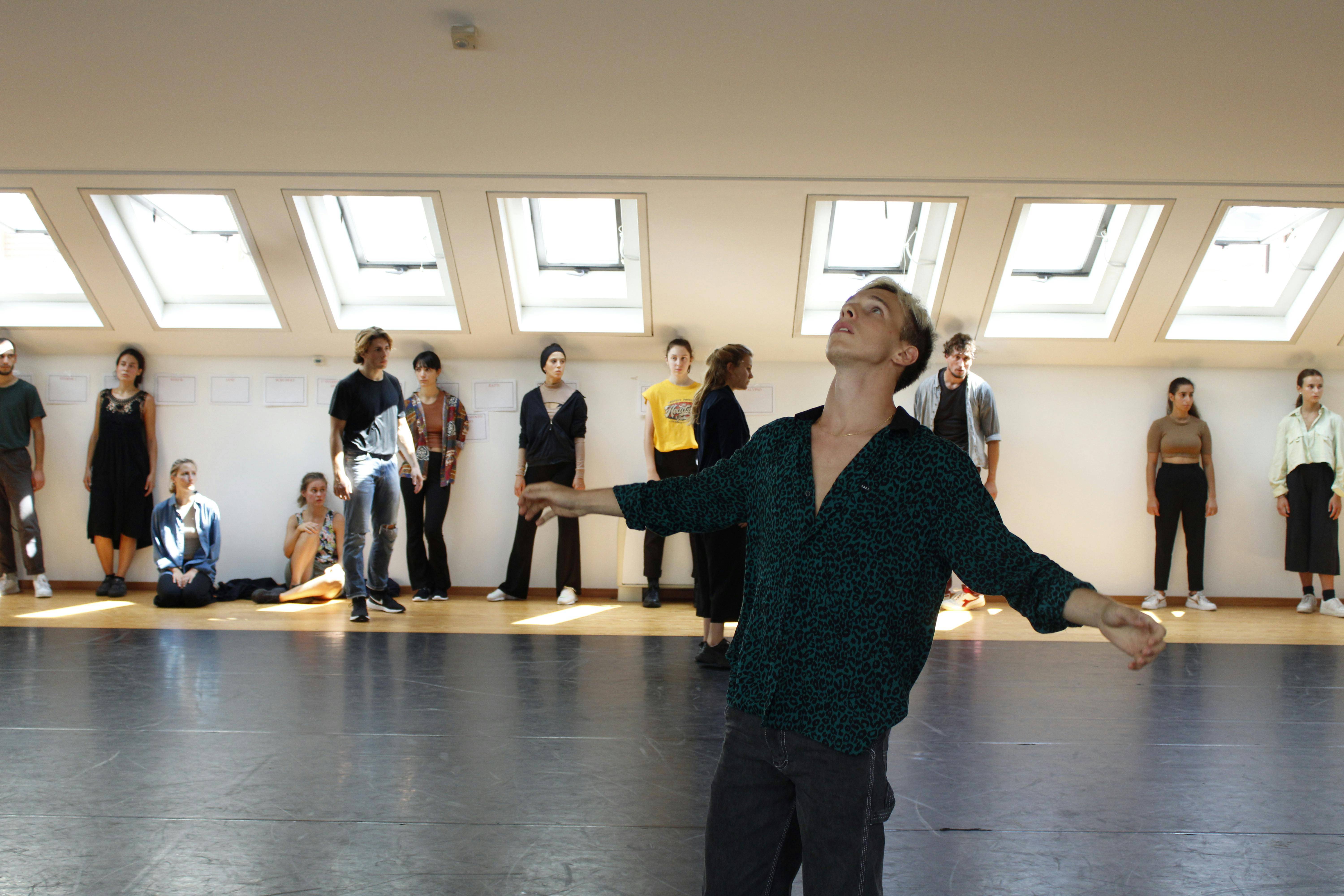 A dancer, standing in the center of the Studio, opens his arms gazing up. At the far end of the room a group of performers, leaning against the wall, observe.
