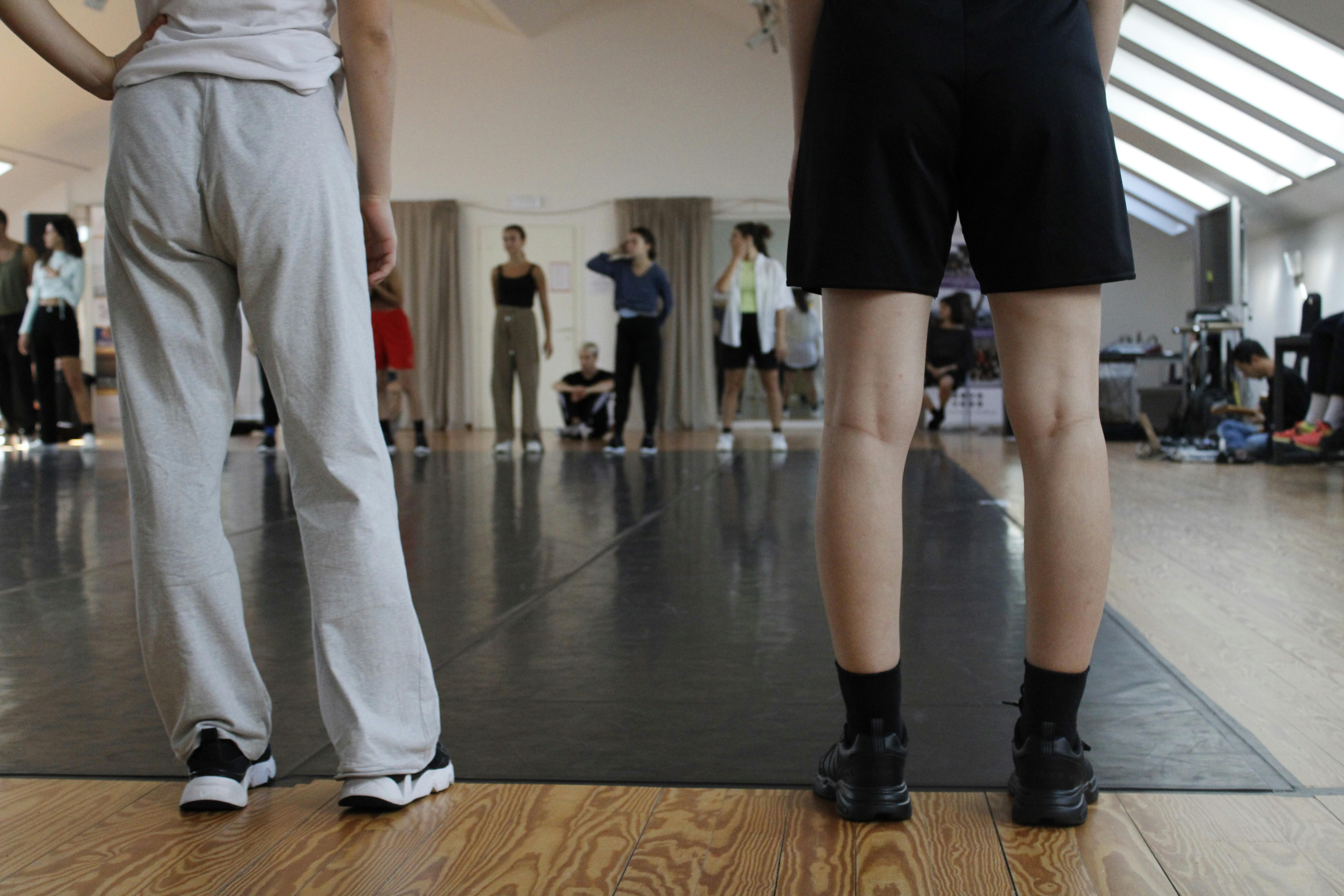 Dancers in residence. In the foreground there are two figures on their backs, visible from the waist down. Opposite, on the other side of the room, other dancers are waiting.