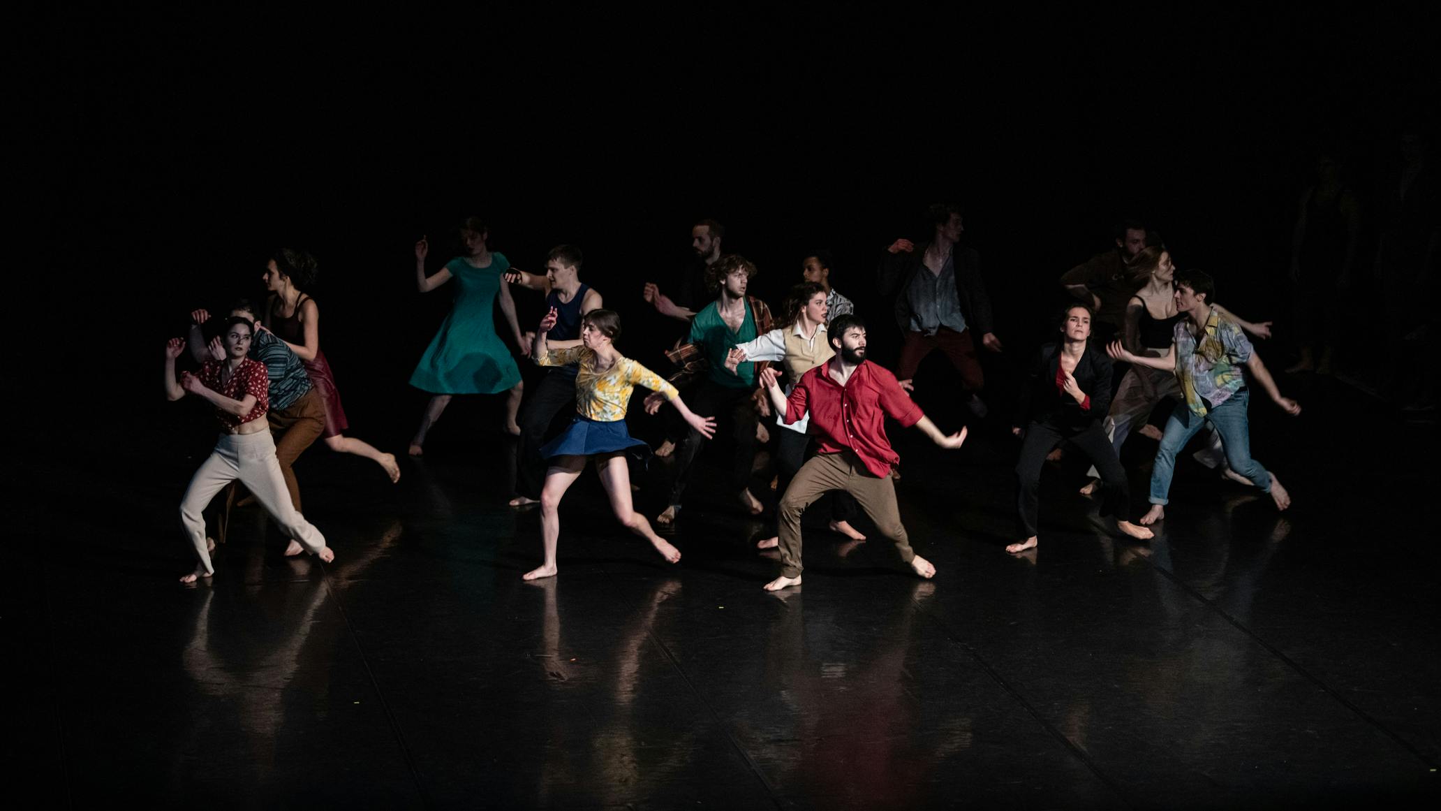 Un gruppo di performer in abiti colorati danza nello spazio. I corpi emergono da sfondo è buio.