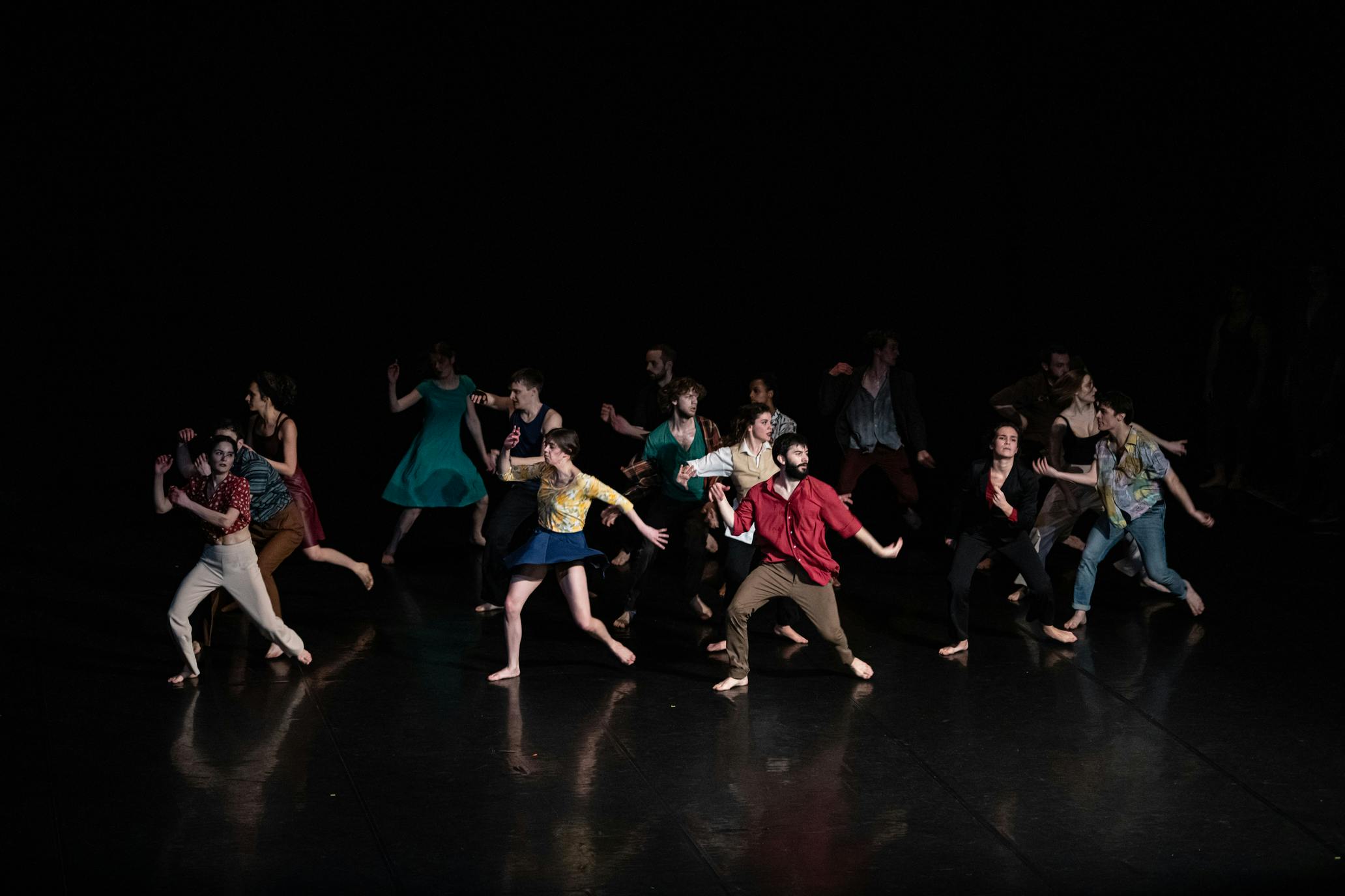 A group of performers in colorful clothing dance in the space. The figures emerge from a dark background.