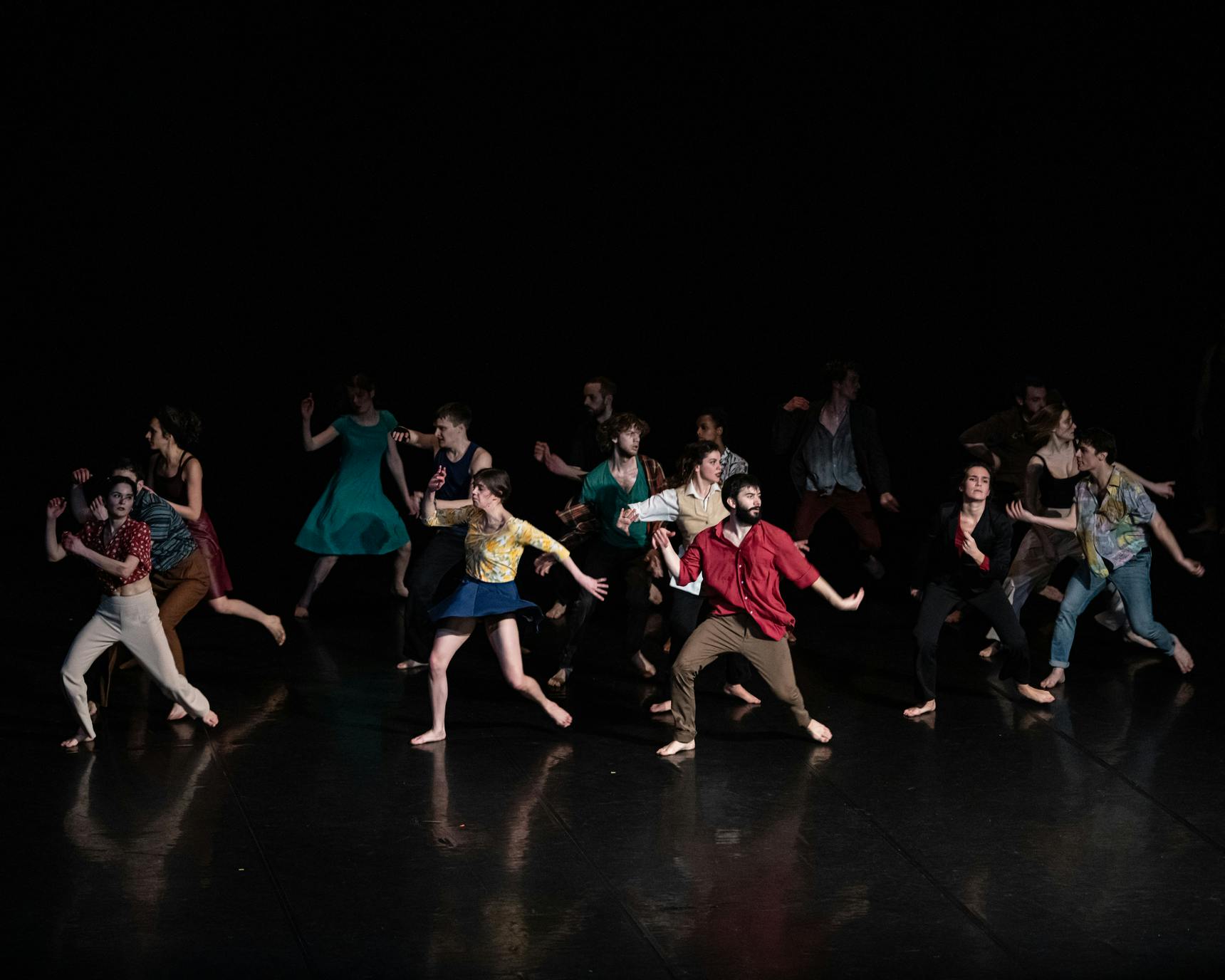 A group of performers in colorful clothing dance in the space. The figures emerge from a dark background.