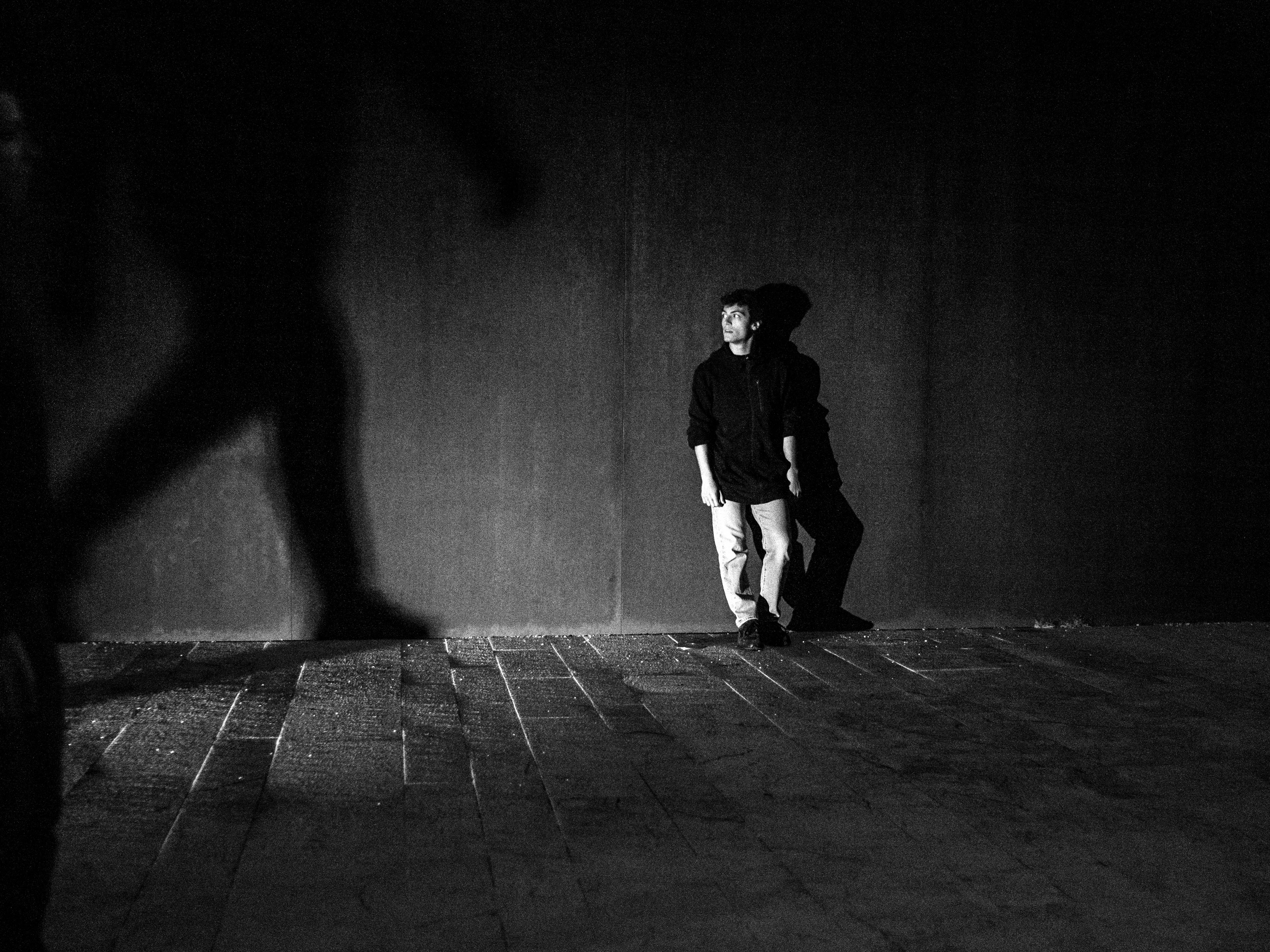 Fotografia in bianco e nero. Un ragazzo, appoggiato ad un muro, è fermo in attesa. Guarda a destra, dove sul proseguo del muro appare grande l'ombra di un corpo che cammina.