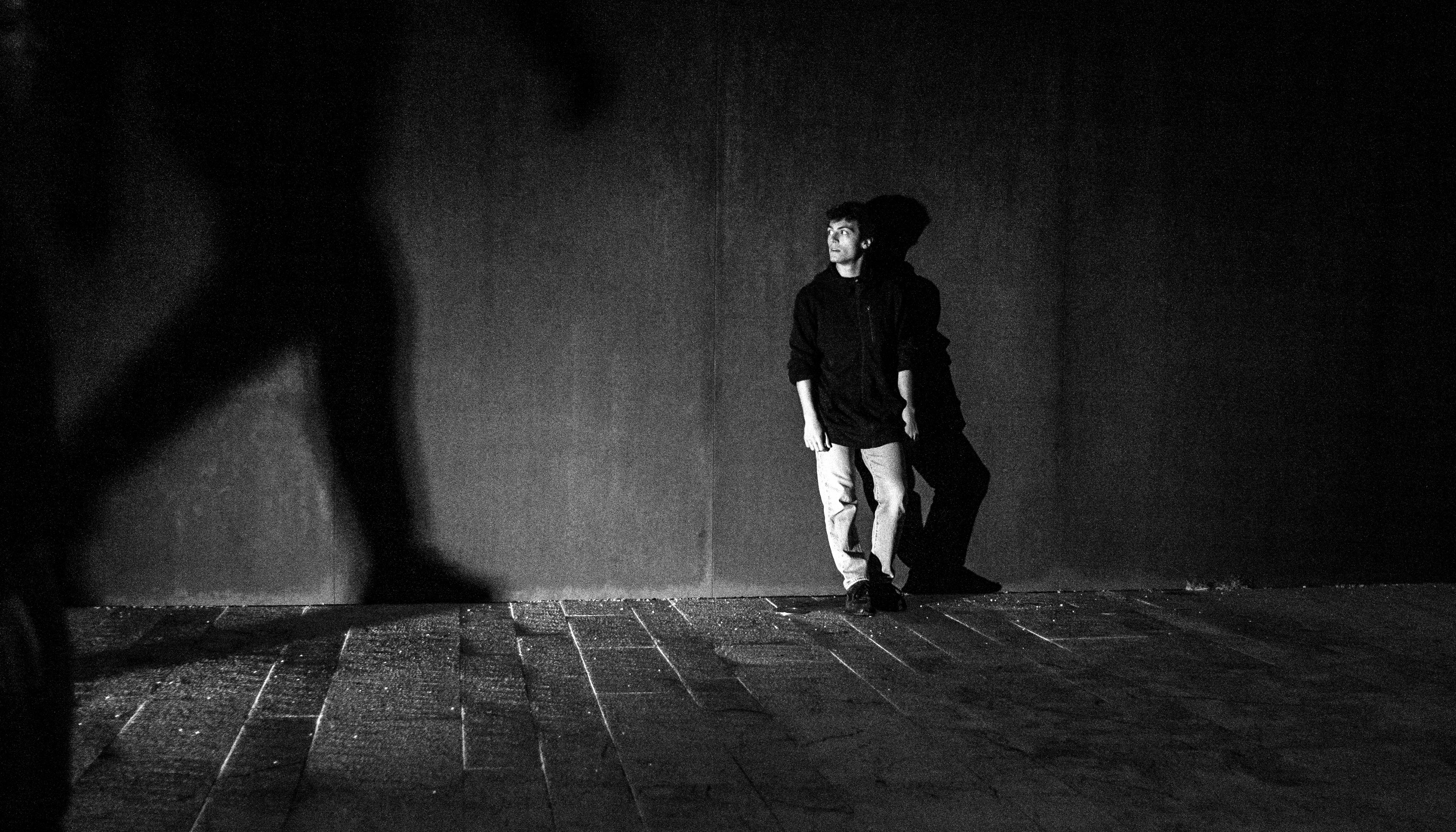 Black and white photograph. A boy, leaning against a wall, is waiting motionless. He looks to the right, where the shadow of a walking body appears large on the wall.