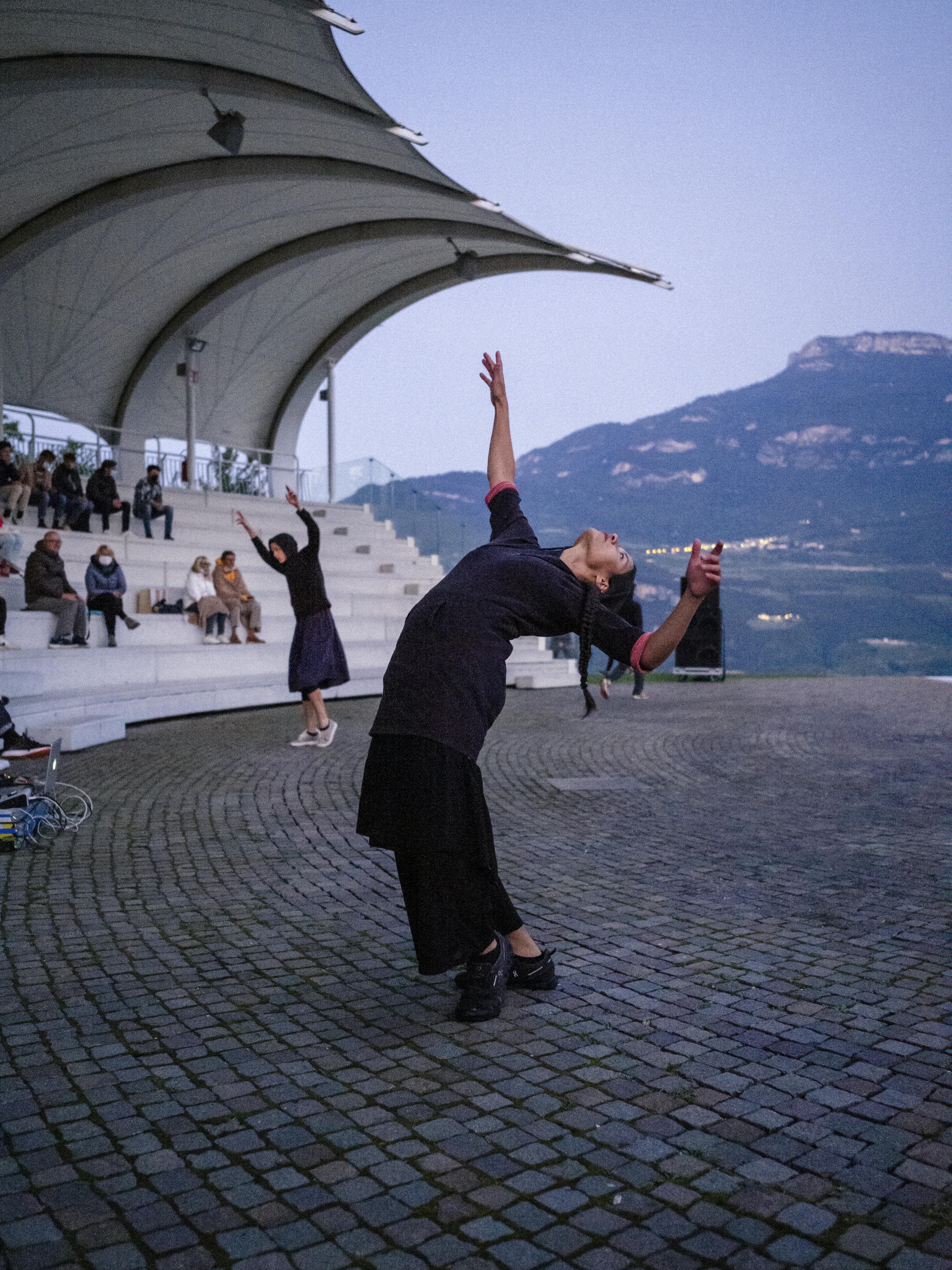 Due danzatrici durante la performance Choros di Alessio Romano alla Campana dei Caduti. La danzatrice in primo piano è in un ampio cambré indietro; braccia e corpo sbilanciati.