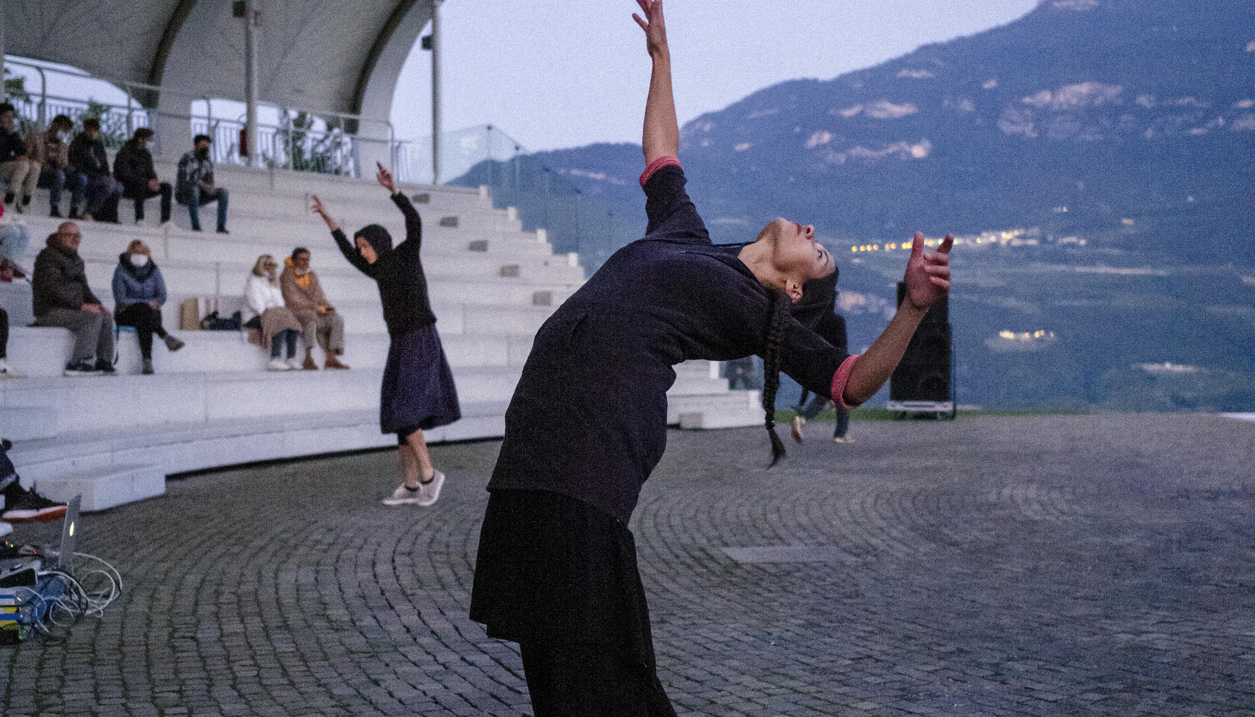 Due danzatrici durante la performance Choros di Alessio Romano alla Campana dei Caduti. La danzatrice in primo piano è in un ampio cambré indietro; braccia e corpo sbilanciati.