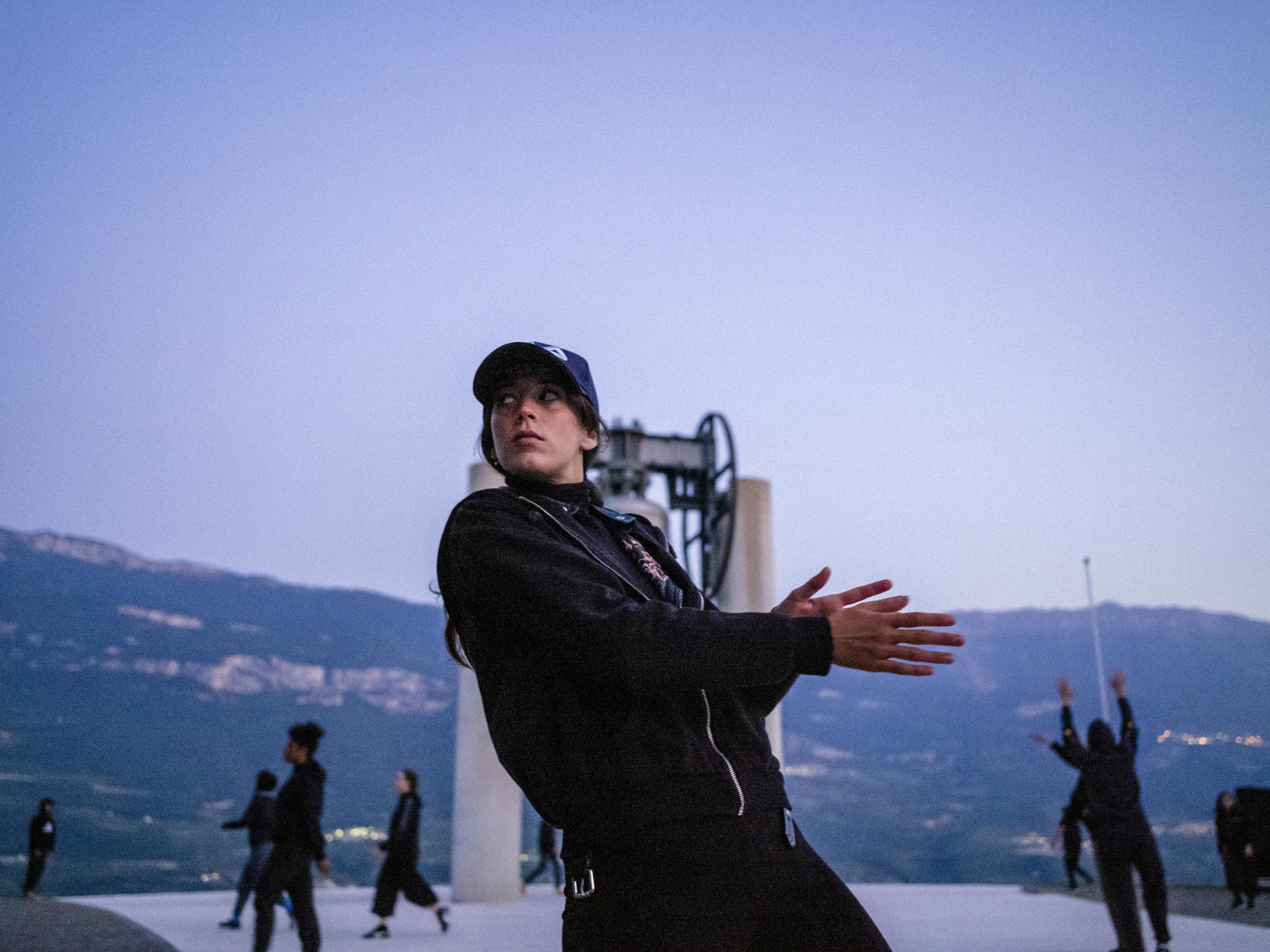 Some performers during Alessio Romano's show "Choros" at the Campana dei Caduti. In the foreground a young dancer dressed in black with a baseball cap. Her body is straight in a diagonal line backwards and she holds her hands palm to palm in front of her body