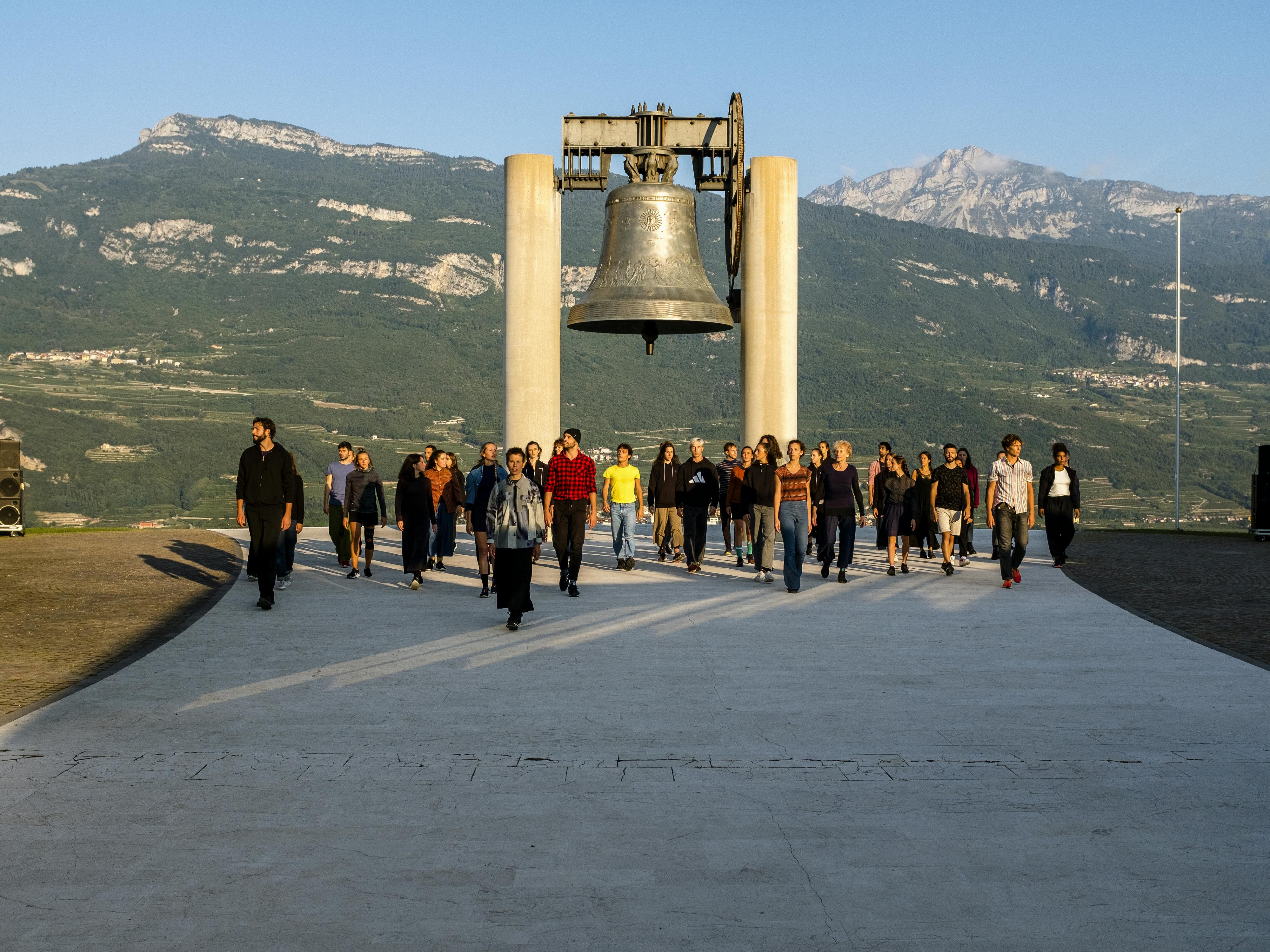 Alessio Maria Romano's performance Choros at the Campana dei Caduti in Rovereto. A large group of performers, in front of the bell, walk towards the camera illuminated by the dawning sun.