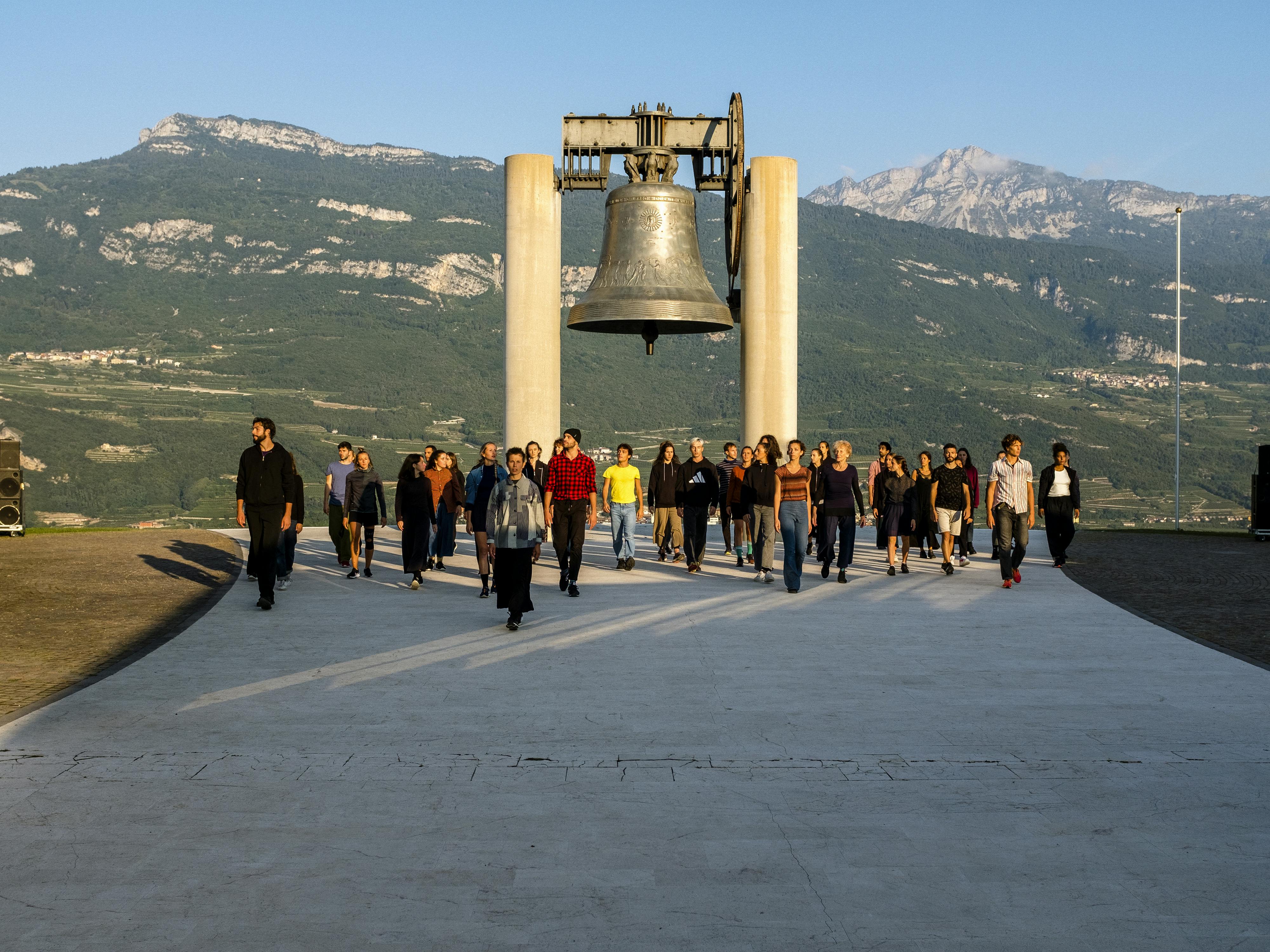 Lo spettacolo Choros di Alessio Maria Romano alla Campana dei Caduti di Rovereto. Un ampio gruppo di performer, davanti alla campana, cammina verso la camera illuminato dal sole dell'alba.