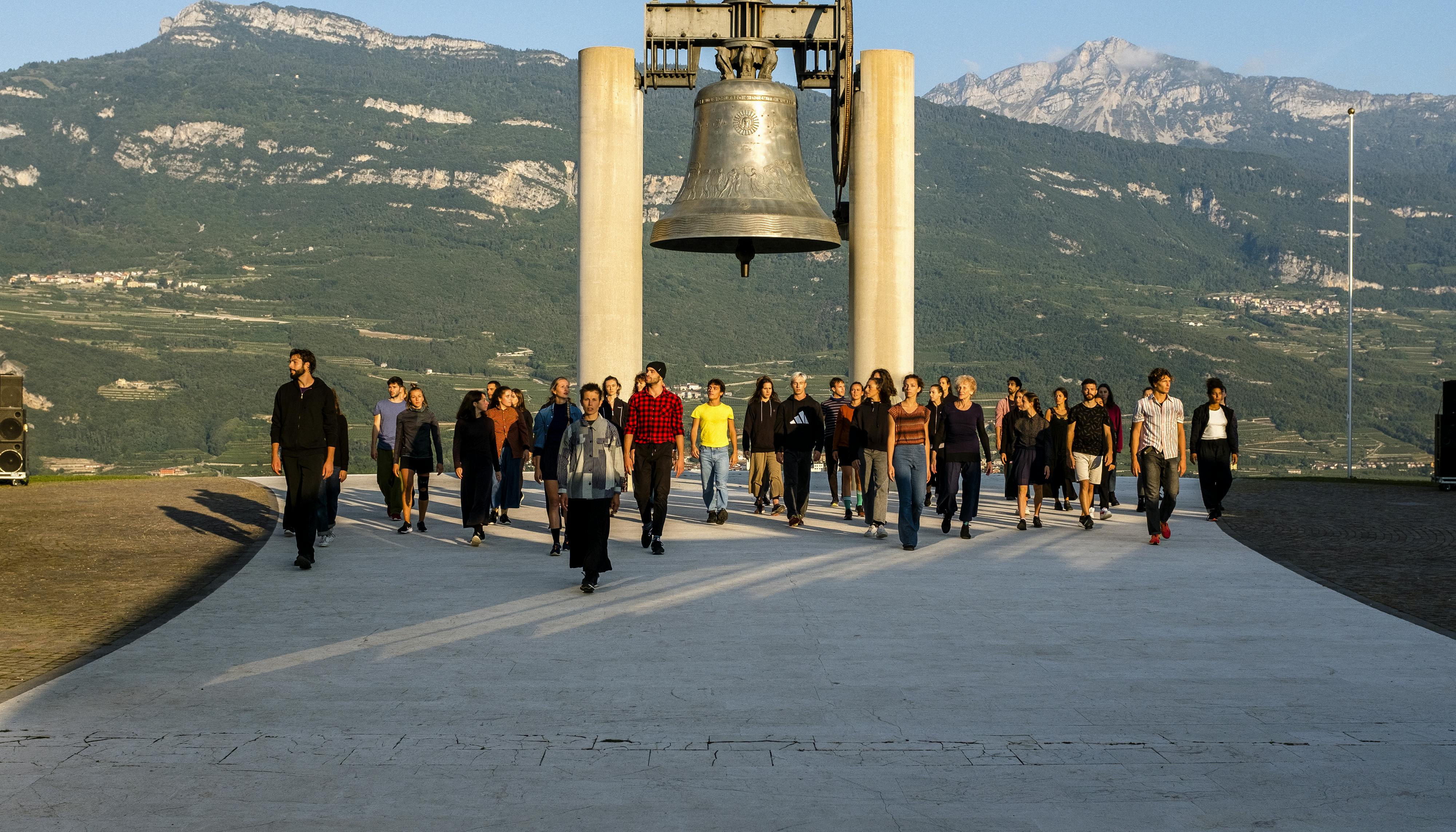 Alessio Maria Romano's performance Choros at the Campana dei Caduti in Rovereto. A large group of performers, in front of the bell, walk towards the camera illuminated by the dawning sun.
