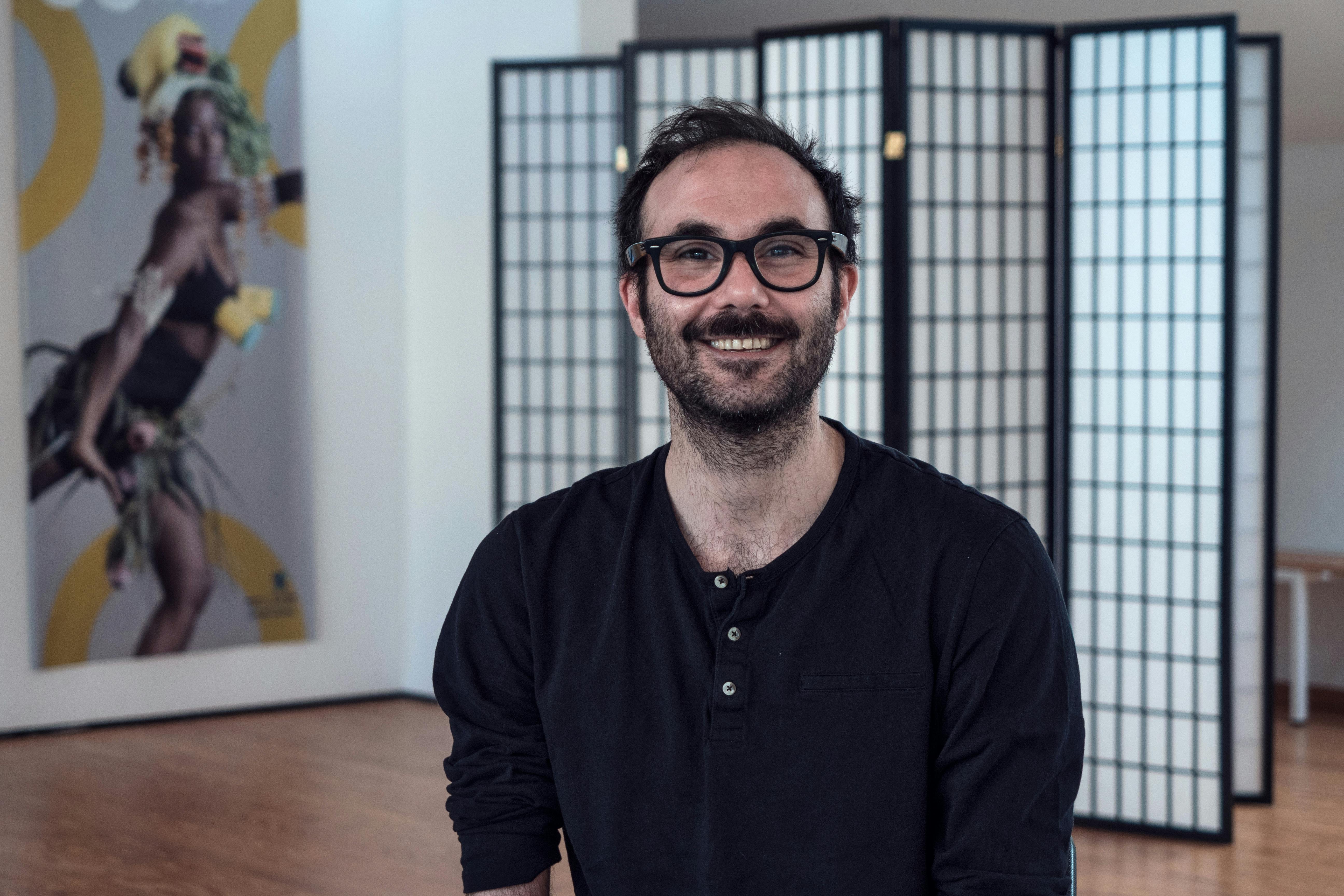 Photo-portrait of Aristide Rontini. He wears a black shirt and black glasses and is smiling at the camera.