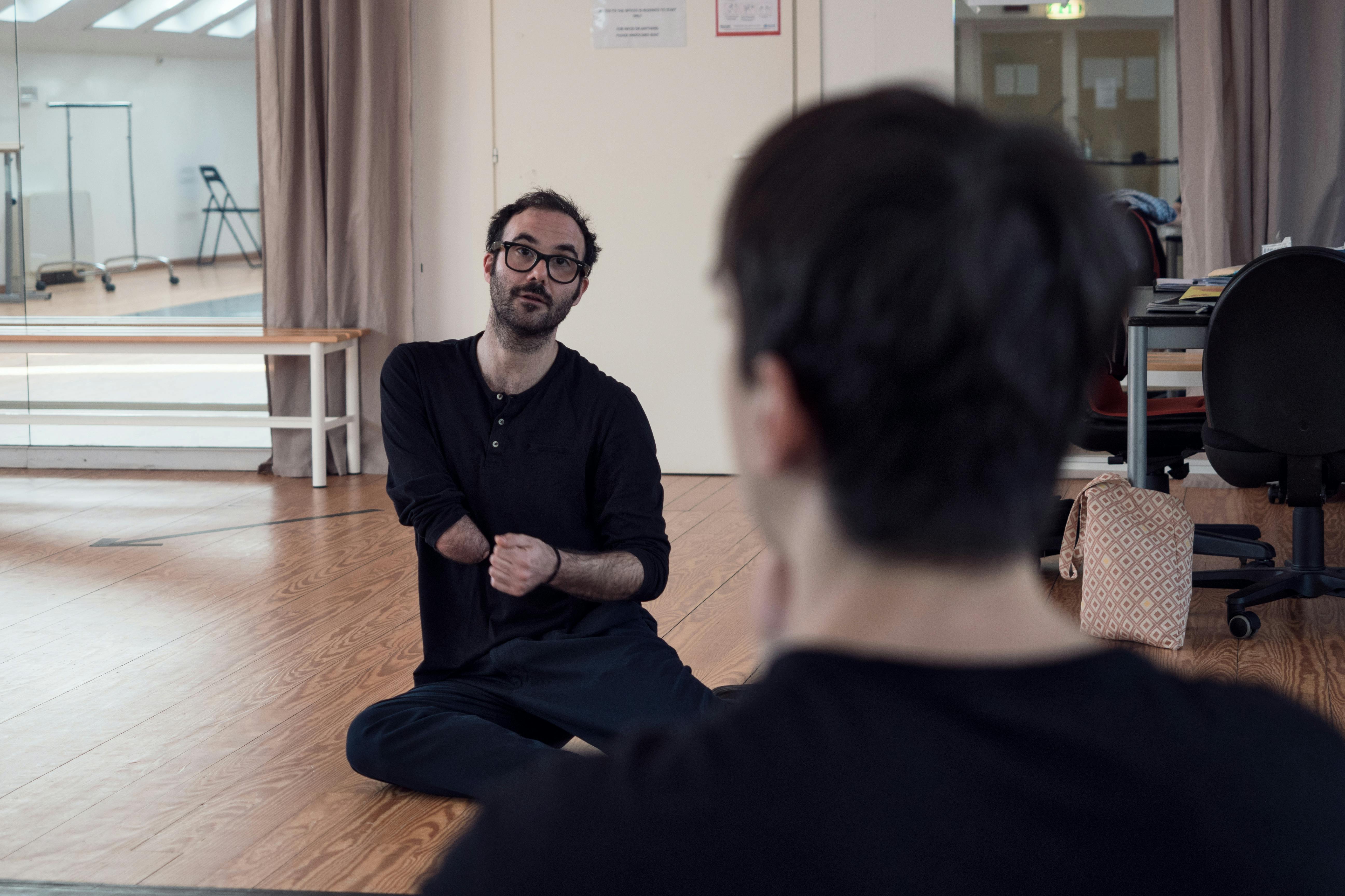 Aristide Rontini, seated on the floor in the Studio, speaks to the dancer Cristian Cucco, seated in front of him.