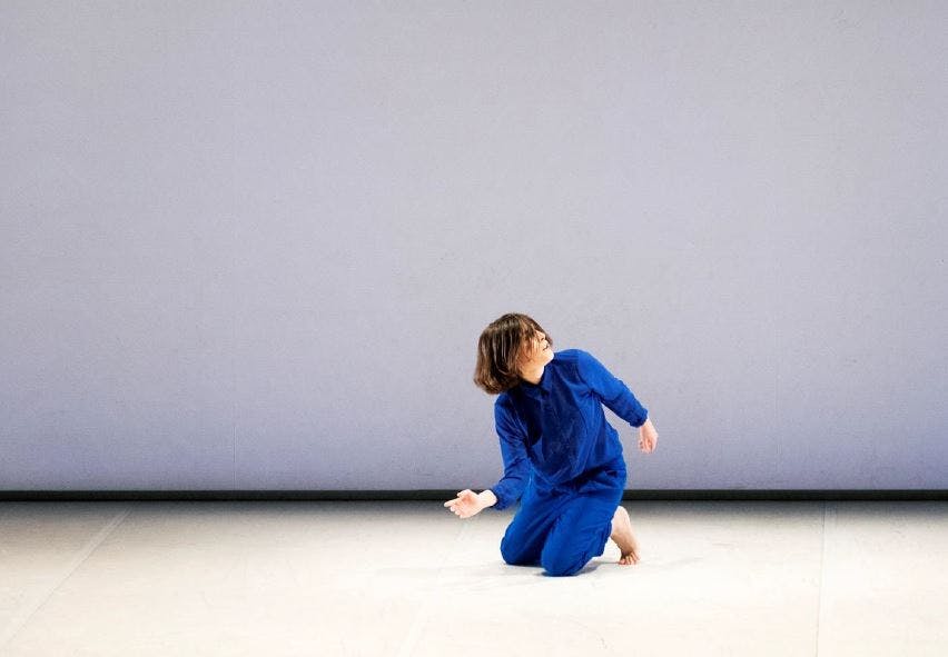 Martina Gambardella in an electric blue jumpsuit, barefoot, while dancing. She is kneeled on the floor, torso and head rotating slightly to the left.