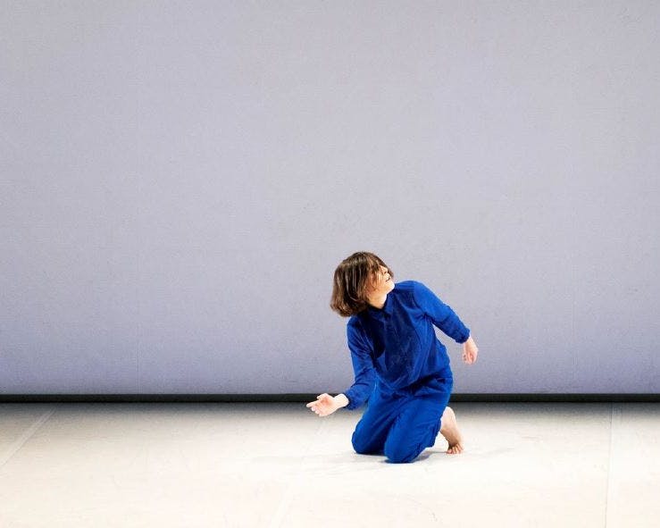 Martina Gambardella in una tuta blu elettrico, a piedi scalzi, mentre danza. È inginocchiata a terra, busto e testa leggermente in rotazione verso sinistra.