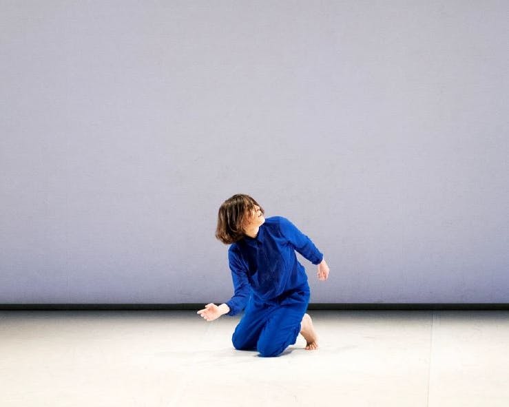 Martina Gambardella in an electric blue jumpsuit, barefoot, while dancing. She is kneeled on the floor, torso and head rotating slightly to the left.
