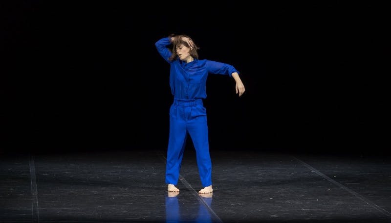 Martina Gambardella in an electric blue jumpsuit, barefoot, as she rehearses Error.  She is standing with her legs slightly apart, one arm raised sideways and the other over her head.