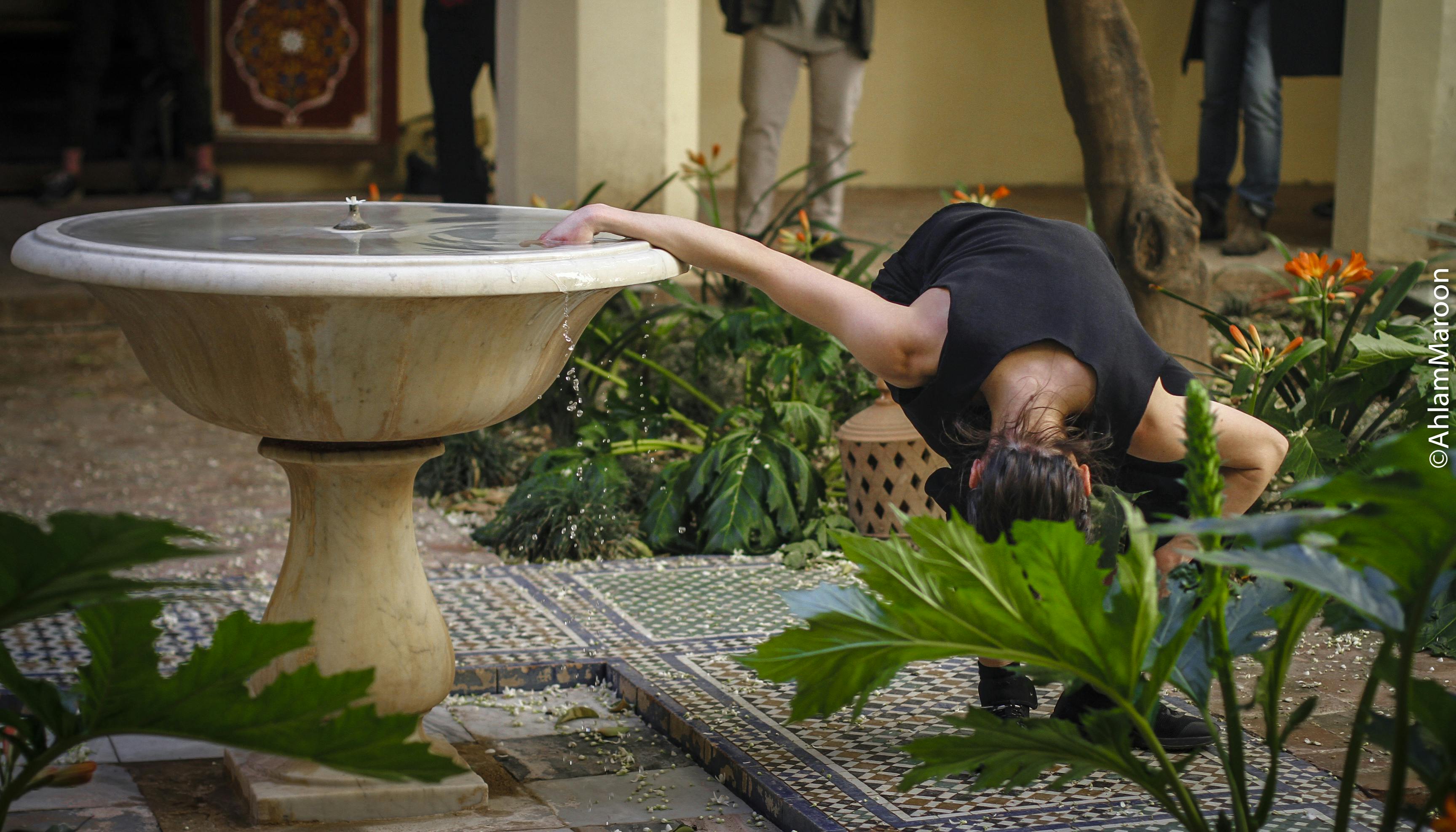 Annamaria Ajmone in an interior courtyard, surrounded by short plants with large leaves and orange flowers. She has one hand inside a fountain while bending with the rest of her body forward, almost to the ground. Behind her, under the colonnade, stands the audience.