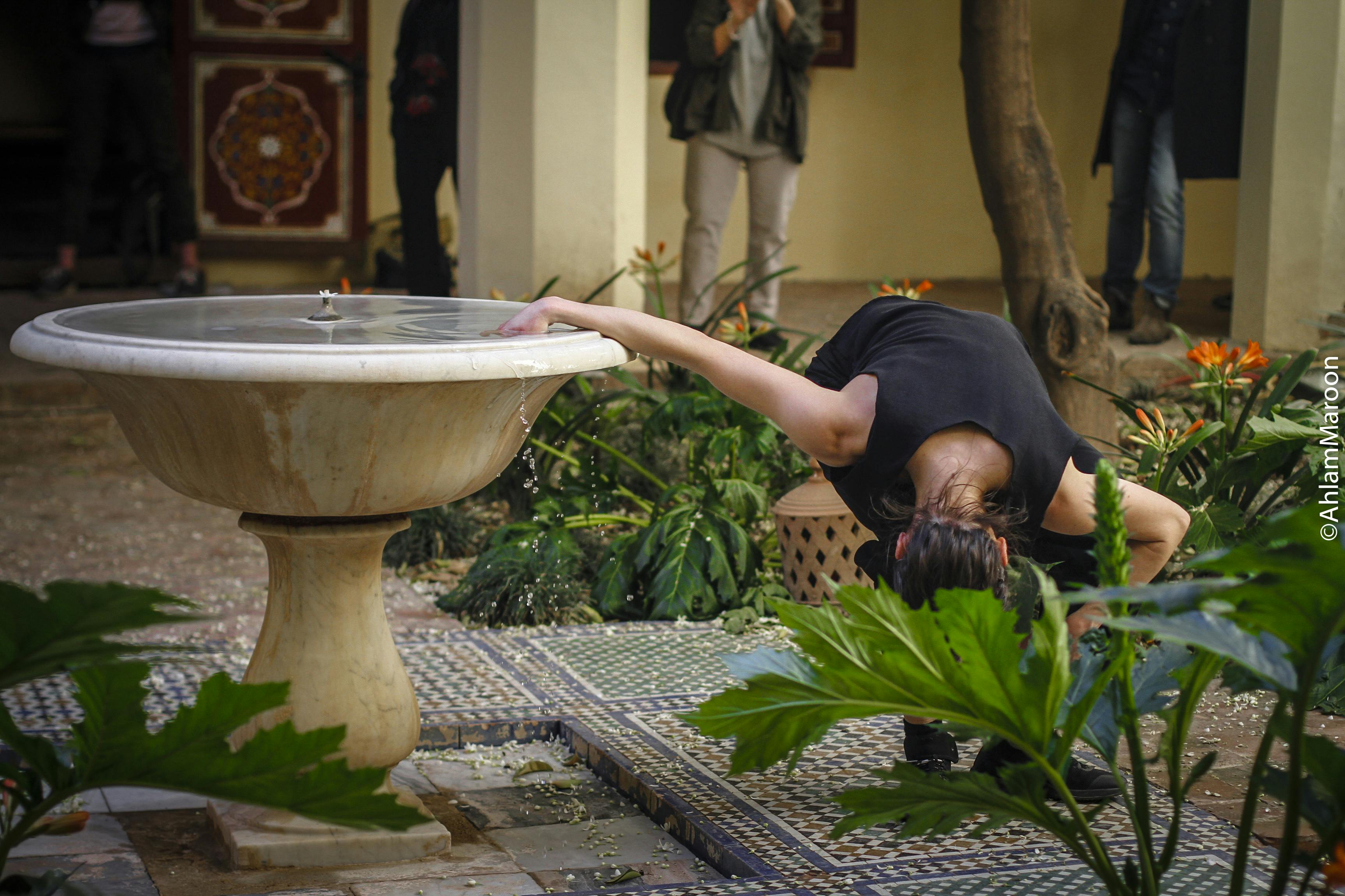 Annamaria Ajmone in an interior courtyard, surrounded by short plants with large leaves and orange flowers. She has one hand inside a fountain while bending with the rest of her body forward, almost to the ground. Behind her, under the colonnade, stands the audience.