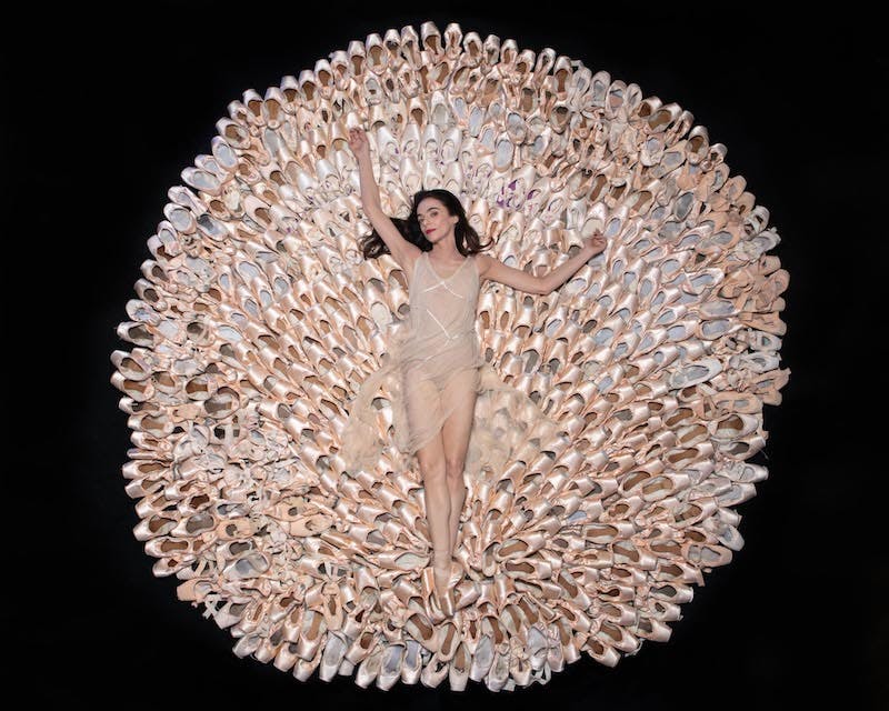 Ballerina Alessandra Ferri is lying on the floor in a light chiffon dress. Below and around her, hundreds of pointe shoes encompass her in a large medallion.