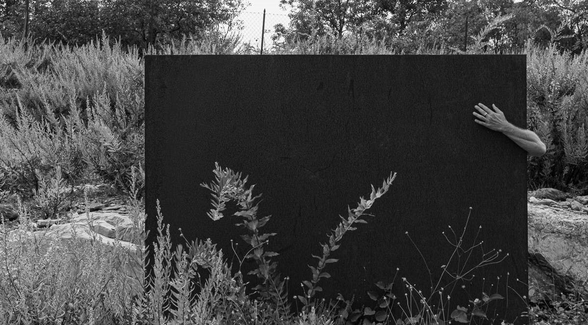 Foto in bianco e nero. Esterno; un grande schermo nero in un giardino. Poggiato su di esso appare da dietro un avanbraccio. Il palmo della mano posa sullo schermo.