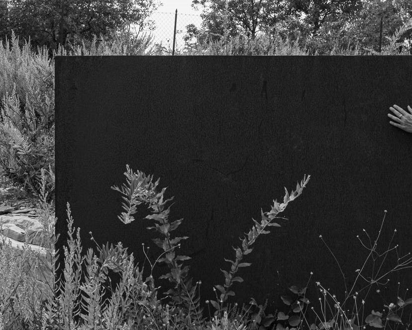 Foto in bianco e nero. Esterno; un grande schermo nero in un giardino. Poggiato su di esso appare da dietro un avanbraccio. Il palmo della mano posa sullo schermo.