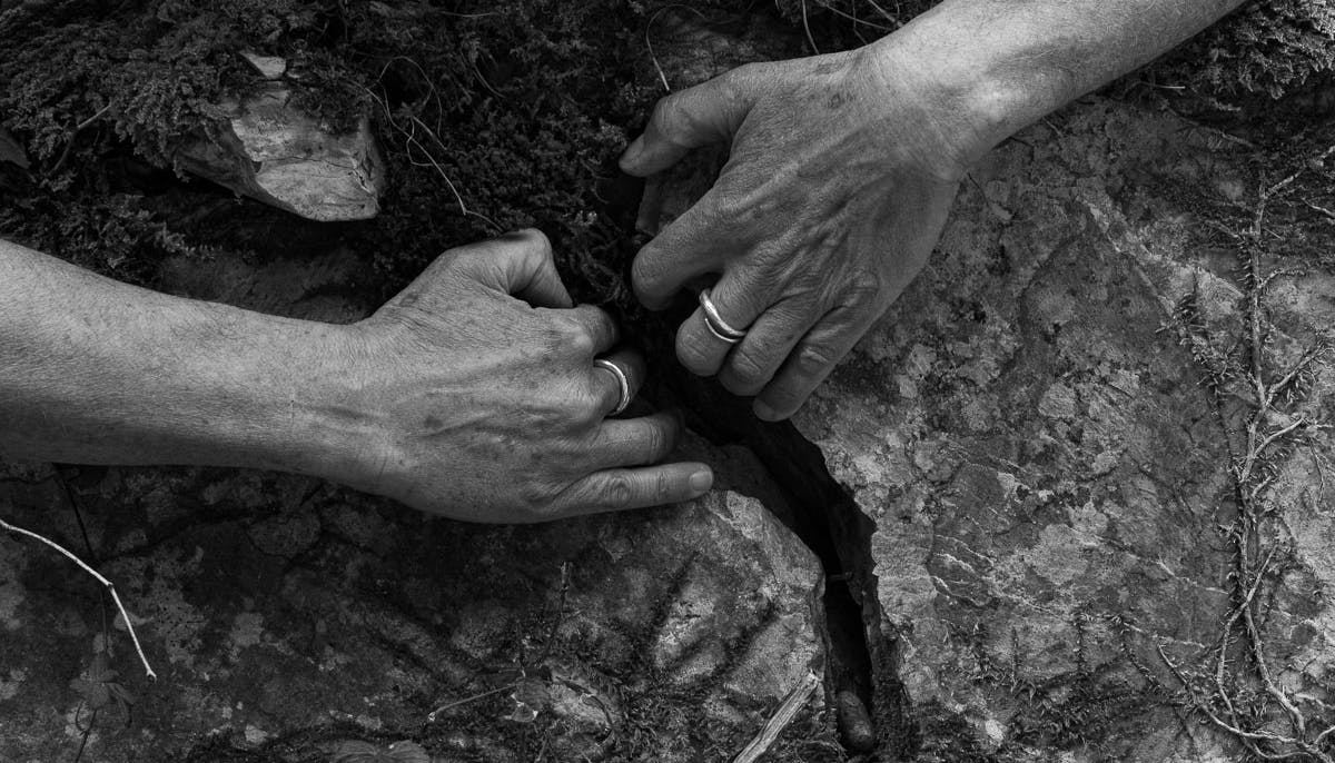 Photo in black and white. Exterior; two hands grasp a crack in the ground pulling as if to widen it.