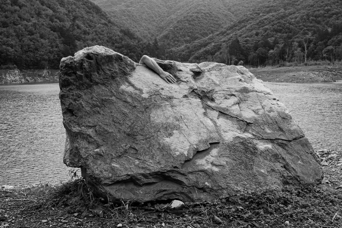 Photo in black and white. Exterior. A large mass in a valley. From above a forearm rests on the stone.