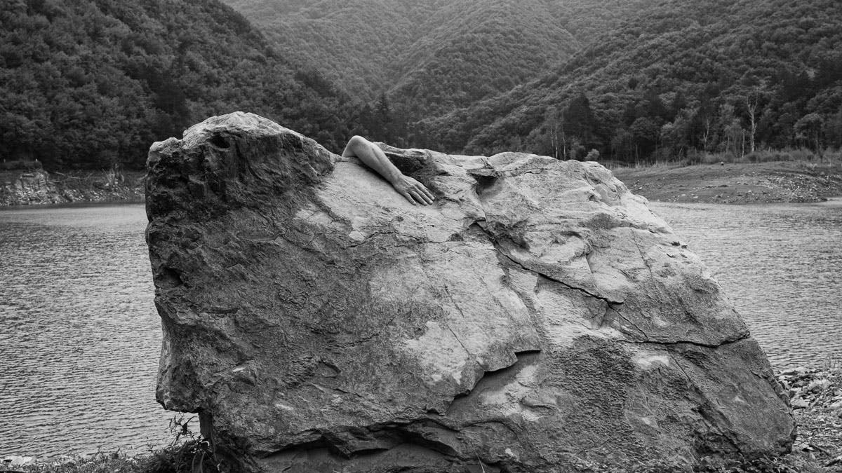 Photo in black and white. Exterior. A large mass in a valley. From above a forearm rests on the stone.