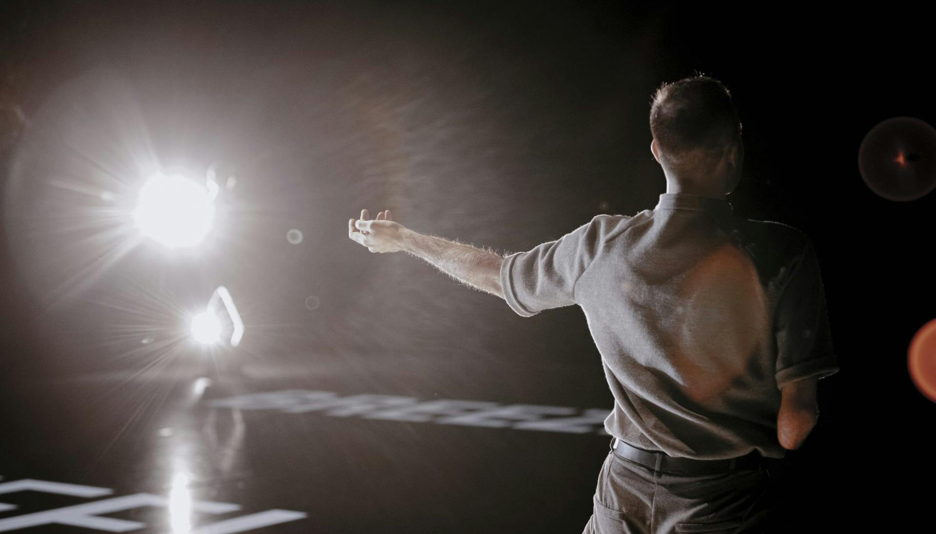 On the stage, from behind, a dancer without a forearm. He is illuminated by a spotlight turned towards the camera.