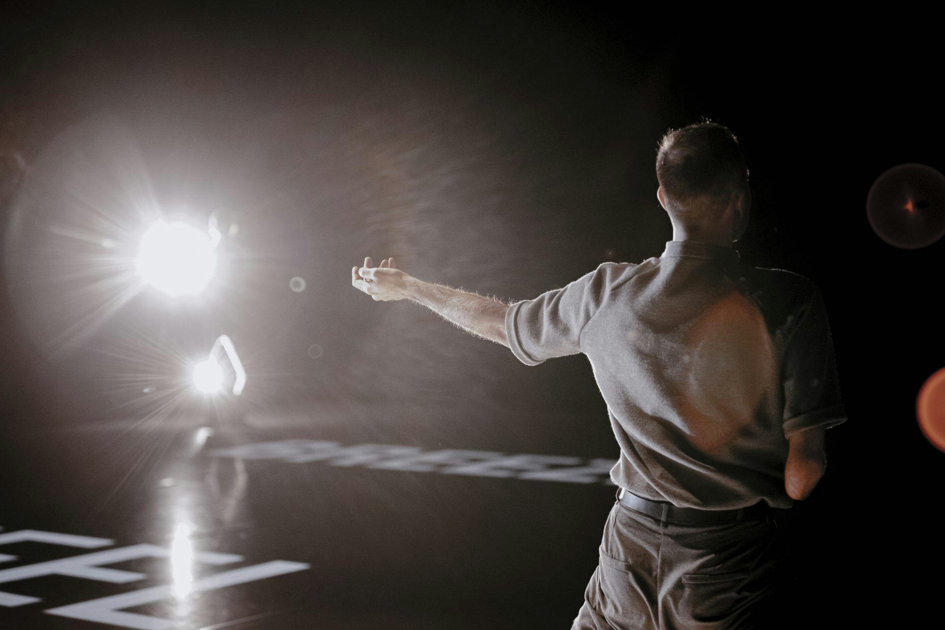 On the stage, from behind, a dancer without a forearm. He is illuminated by a spotlight turned towards the camera.