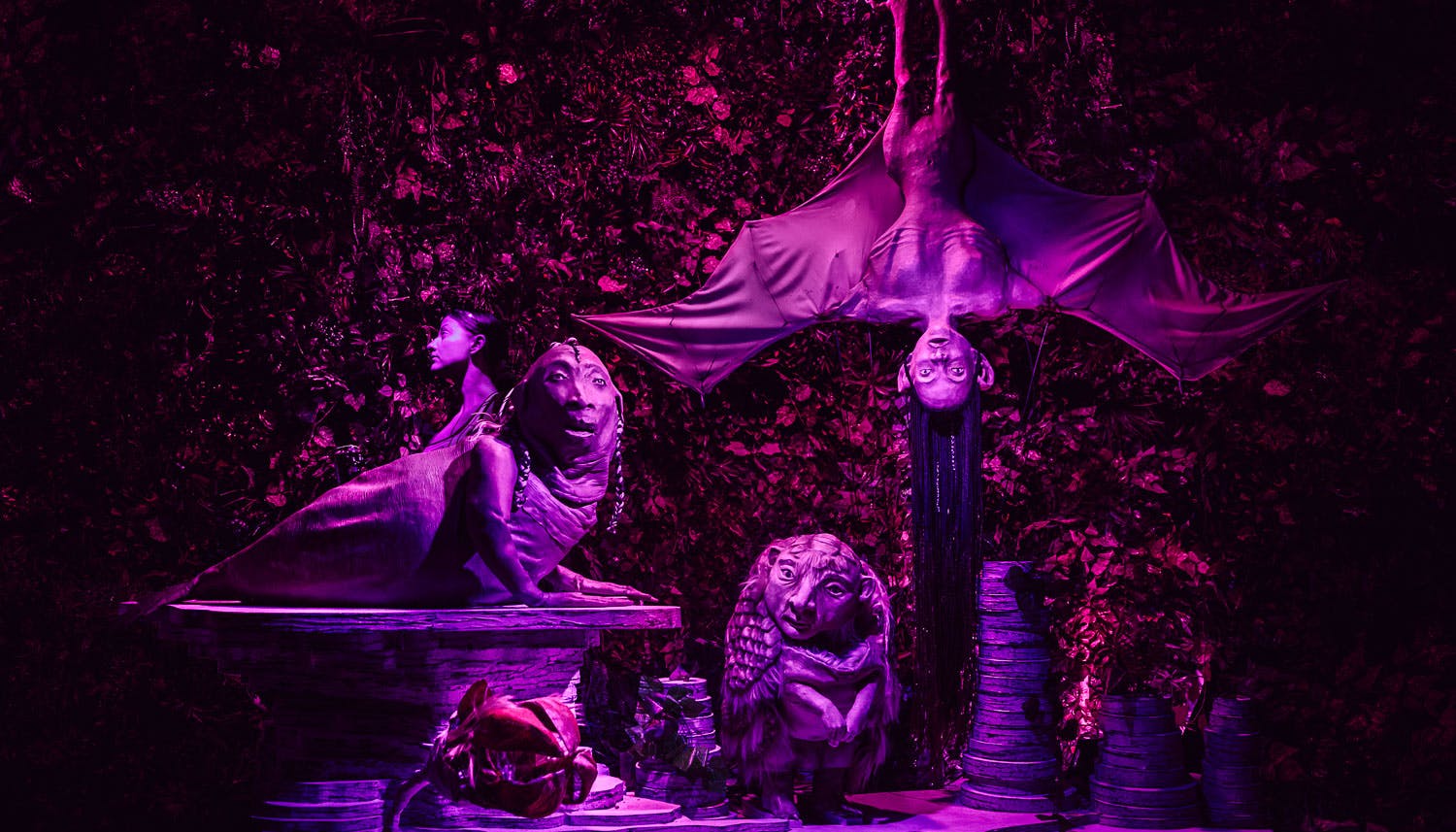 A surreal scenography. Strange sculptures with figures partly zoomorphic and partly human, in front of a background of foliage. Everything is illuminated by a soft violet light.