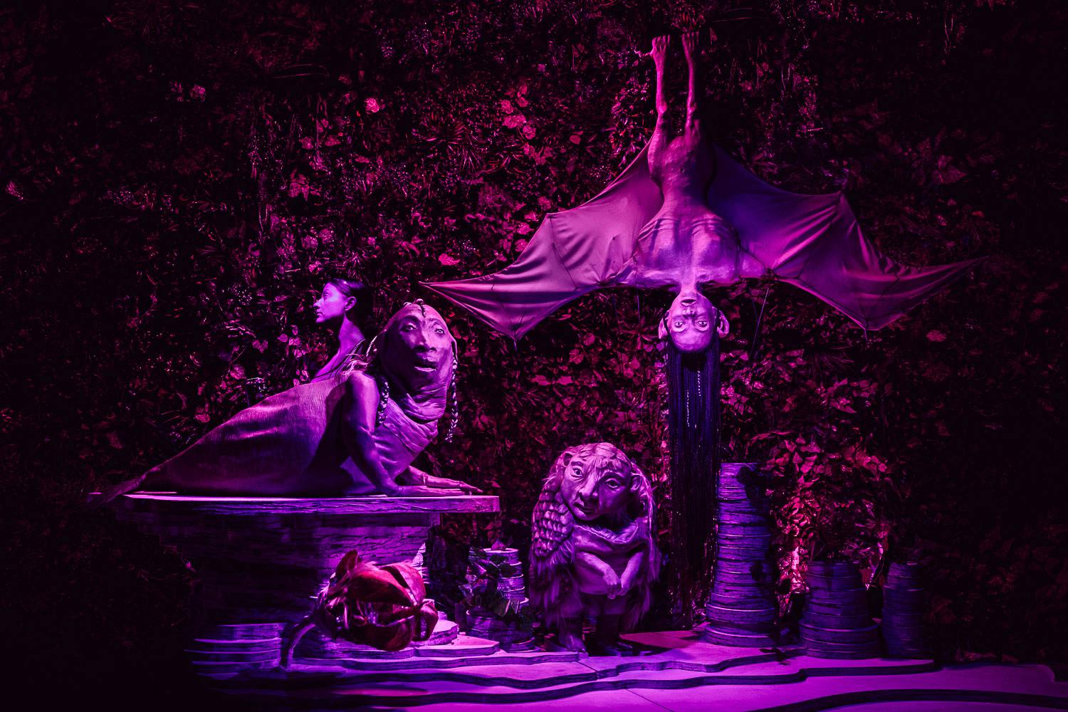 A surreal scenography. Strange sculptures with figures partly zoomorphic and partly human, in front of a background of foliage. Everything is illuminated by a soft violet light.