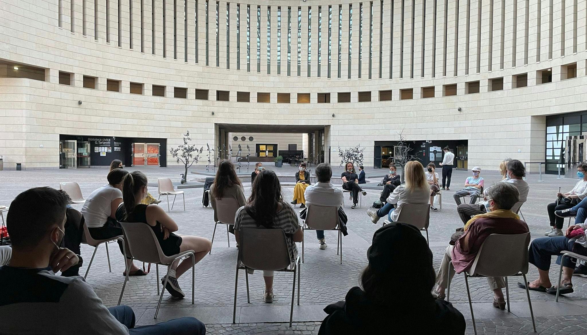 Listening point for "Dimmi cosa vuoi vedere" in the Mart square in Rovereto. The audience is visible from the back; facing them Marta Cuscunà and Al.Di.Qua.Artist are seated in a semicircle.