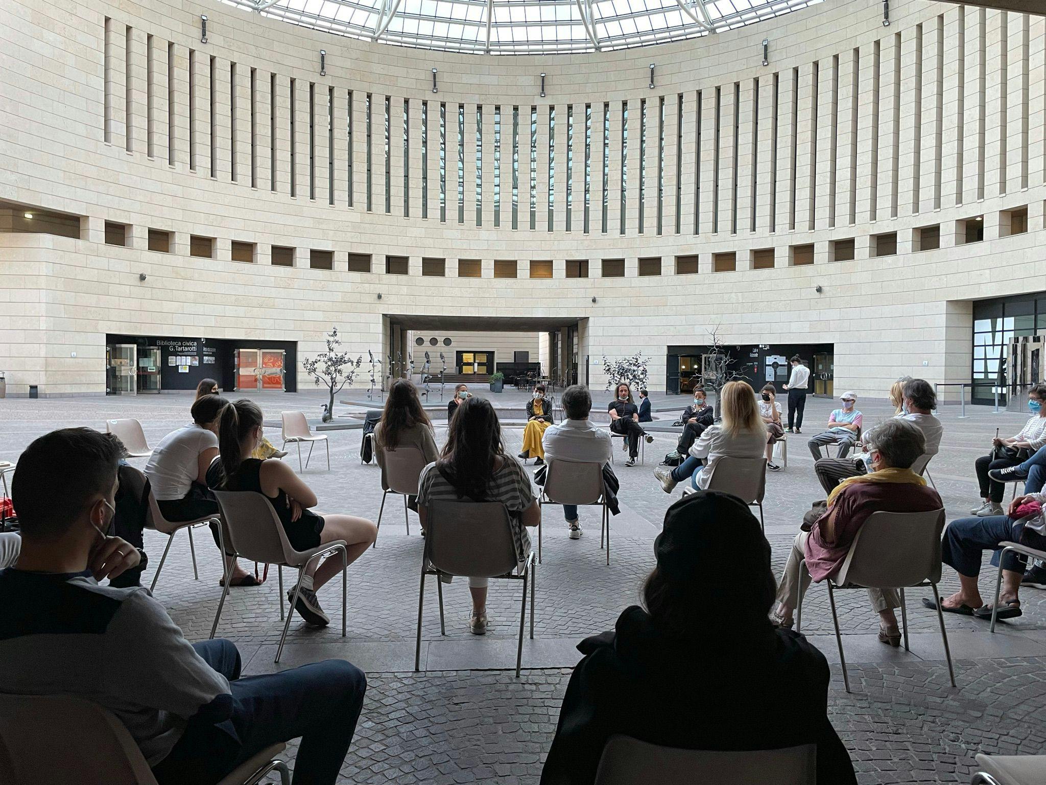 Punto di ascolto di "Dimmi cosa vuoi vedere" nella piazza del Mart di Rovereto. Il pubblico è visibile di schiena; di fronte Marta Cuscunà e Al.Di.Qua.Artist sono seduti a semicerchio.