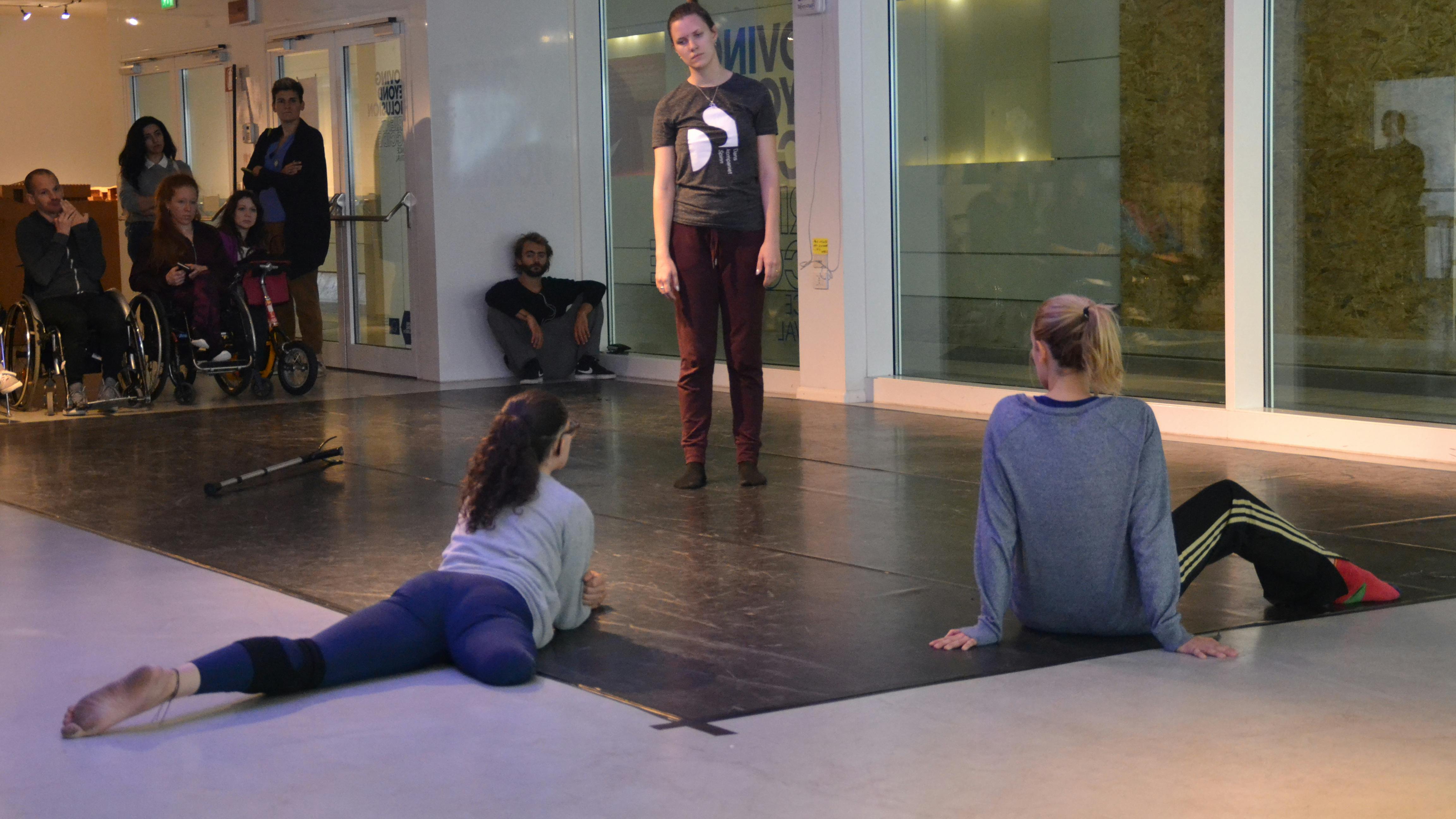 Three dancers, one of them without a leg, during a workshop. Other participants with and without disabilities observe from the back of the room.