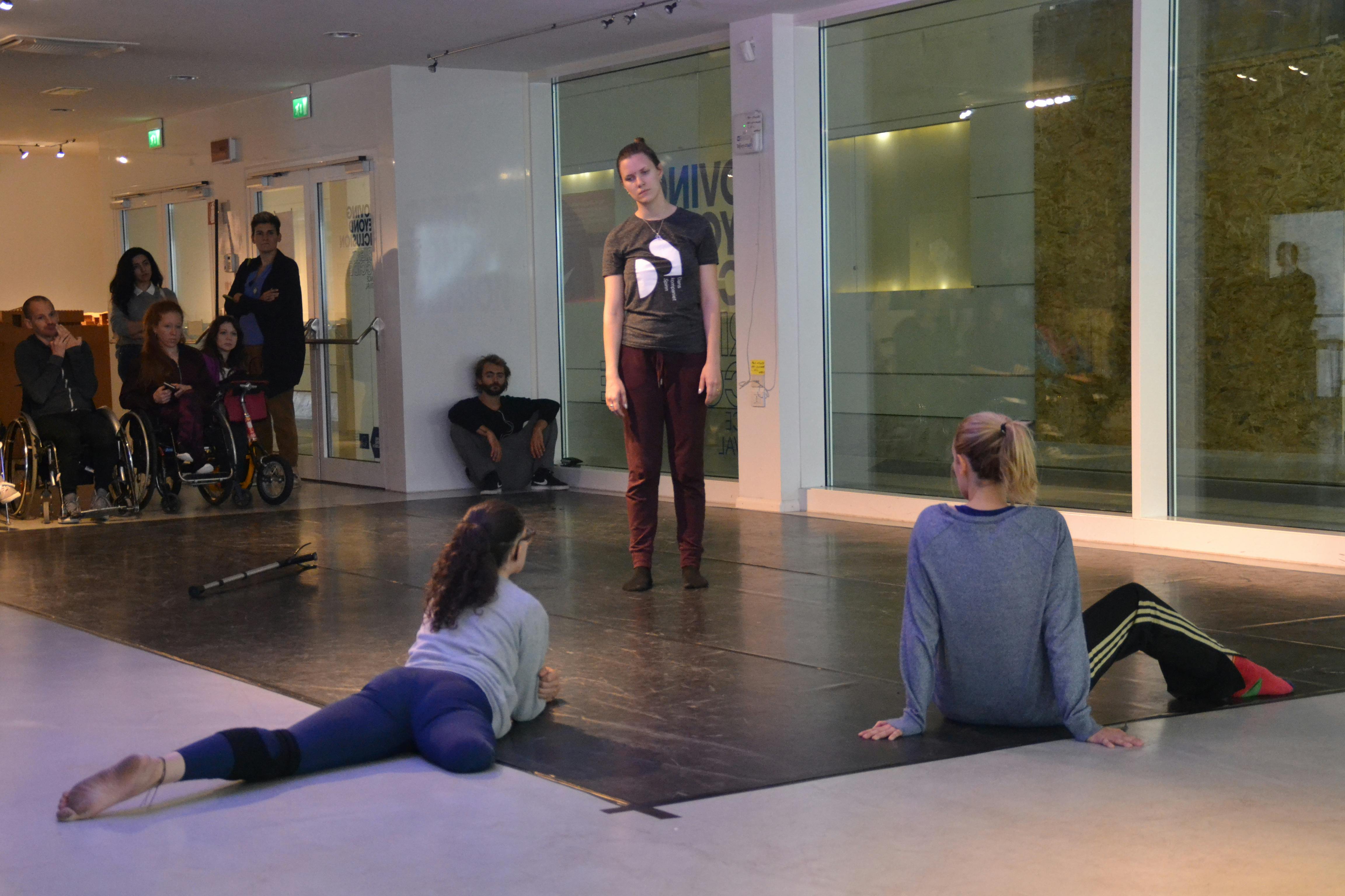Three dancers, one of them without a leg, during a workshop. Other participants with and without disabilities observe from the back of the room.