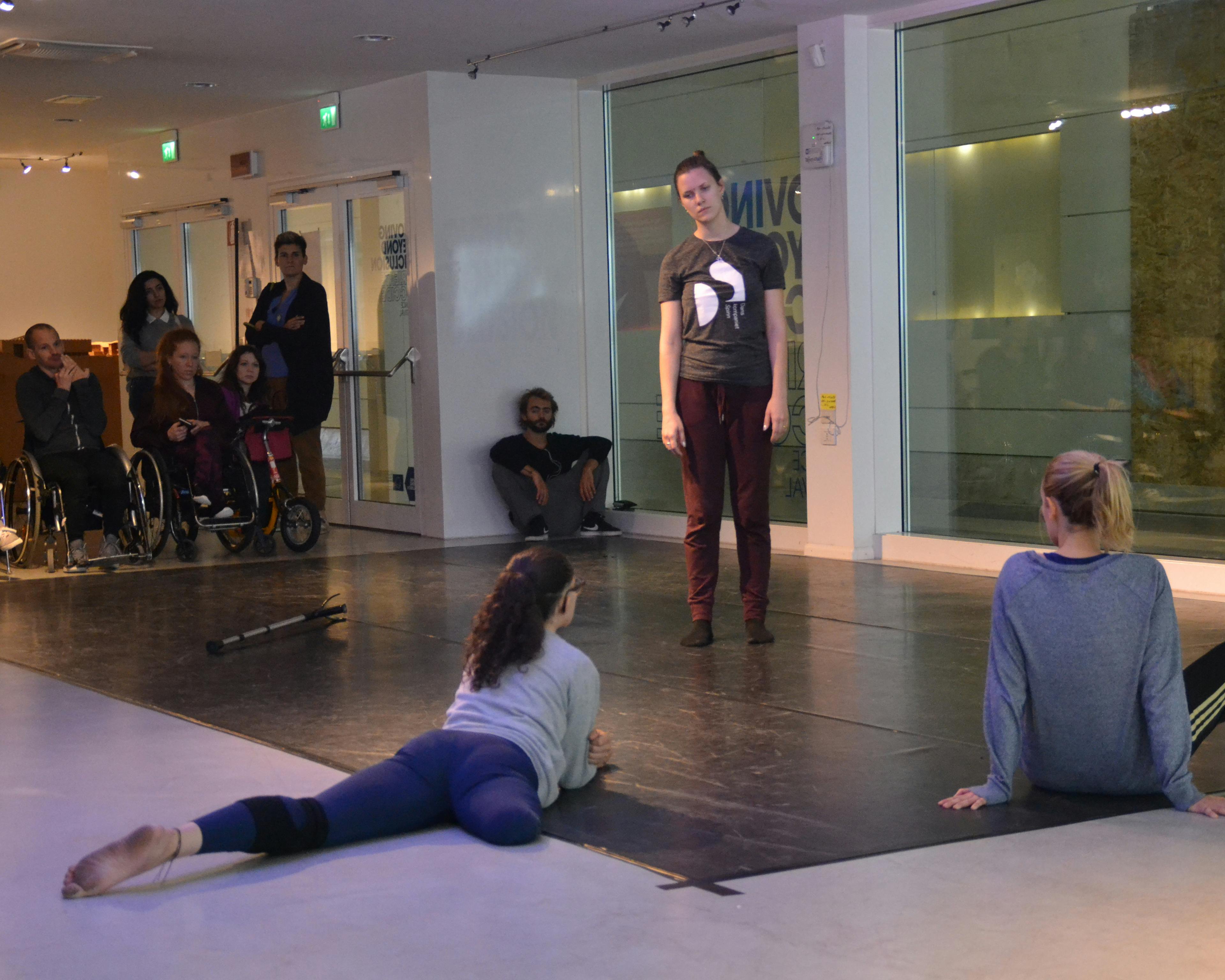 Tre danzatrici, una delle quali senza una gamba, durante un laboratorio. Altre ed altri partecipanti con e senza disabilità osservano da fondo sala.