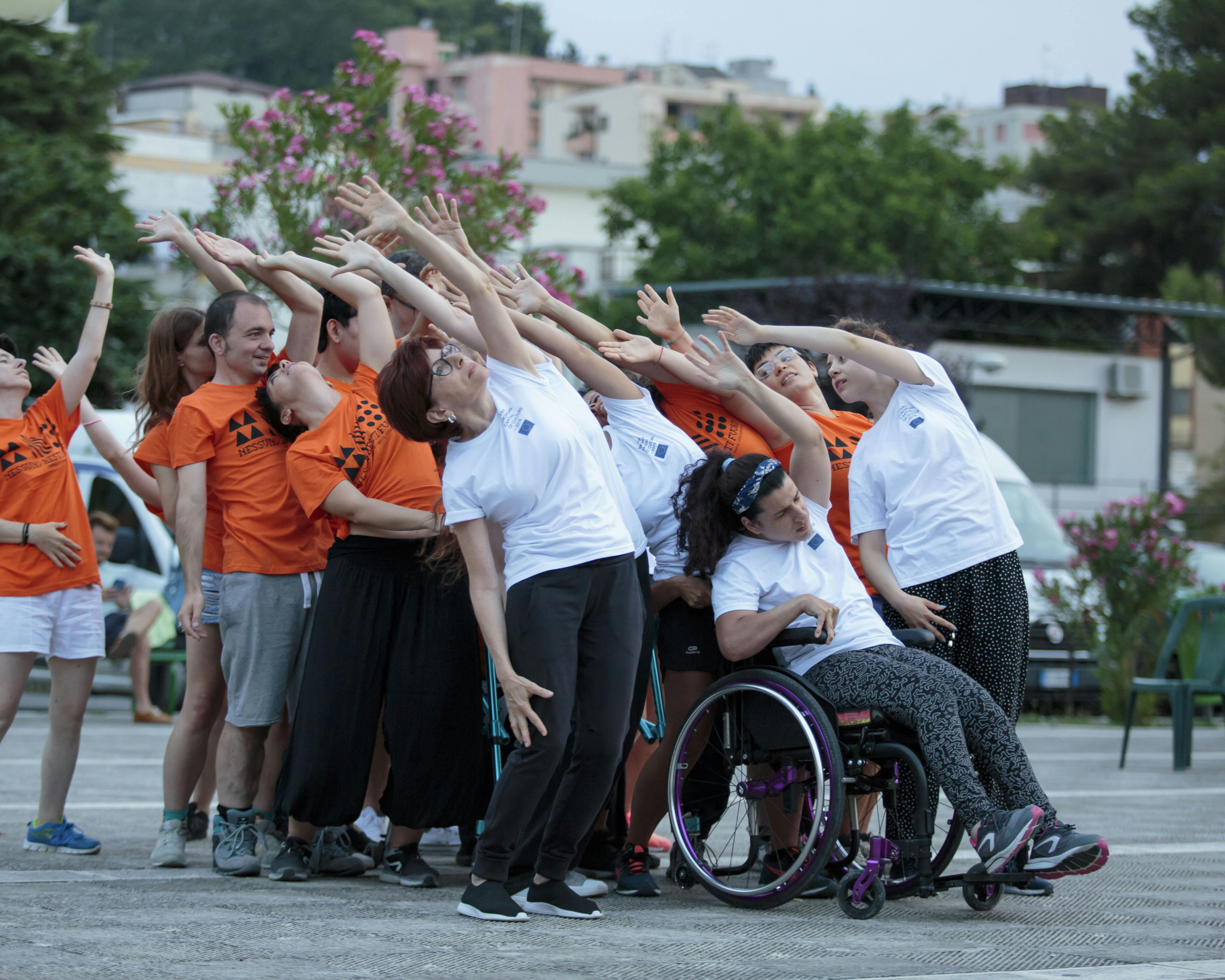 A group of performers with and without disabilities during a performance. They raise one arm upward while slightly tilting their torso back.